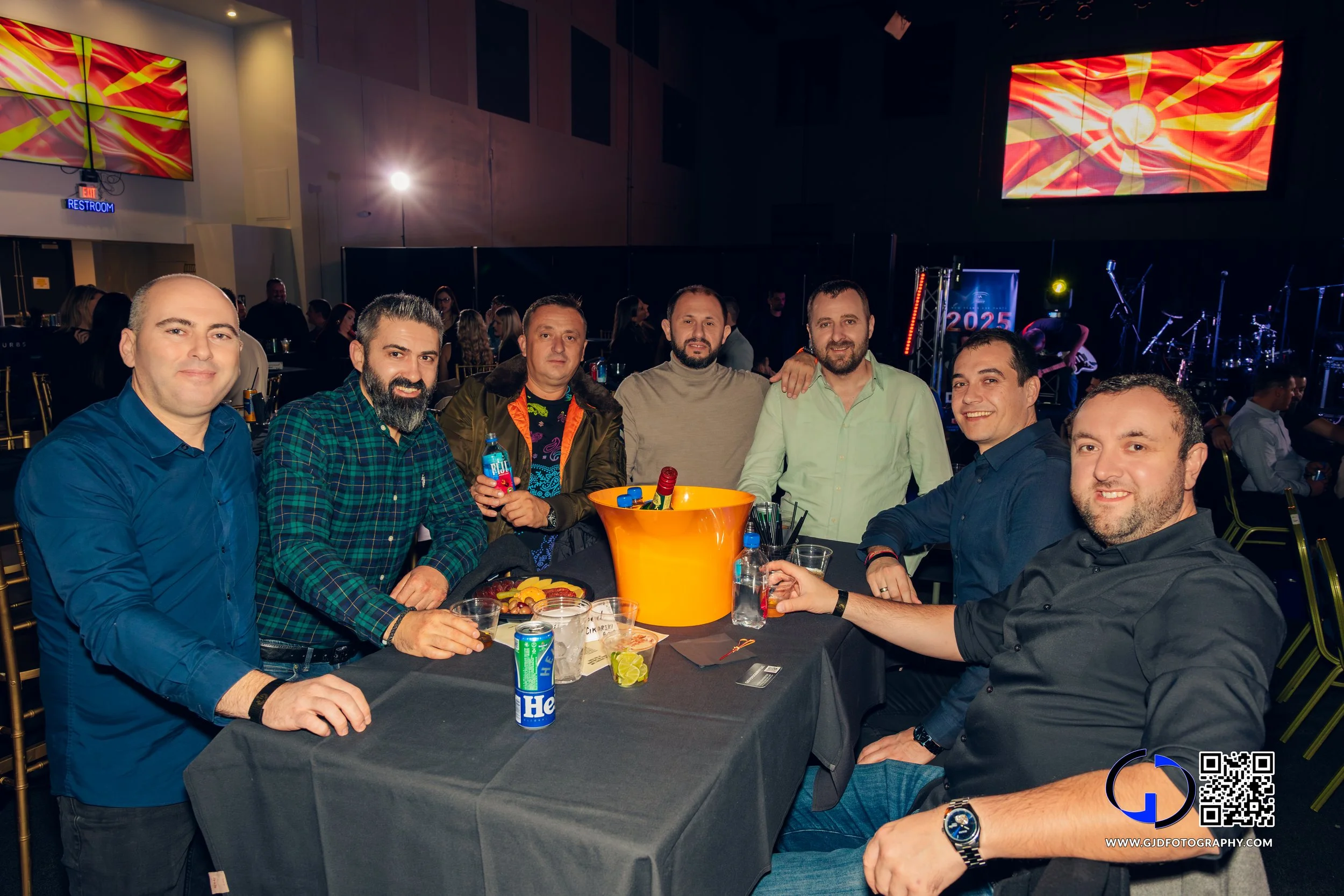 Group of seven men sitting around a table at a social event, with drinks and snacks, in a dimly lit venue with a stage and large screen in the background.