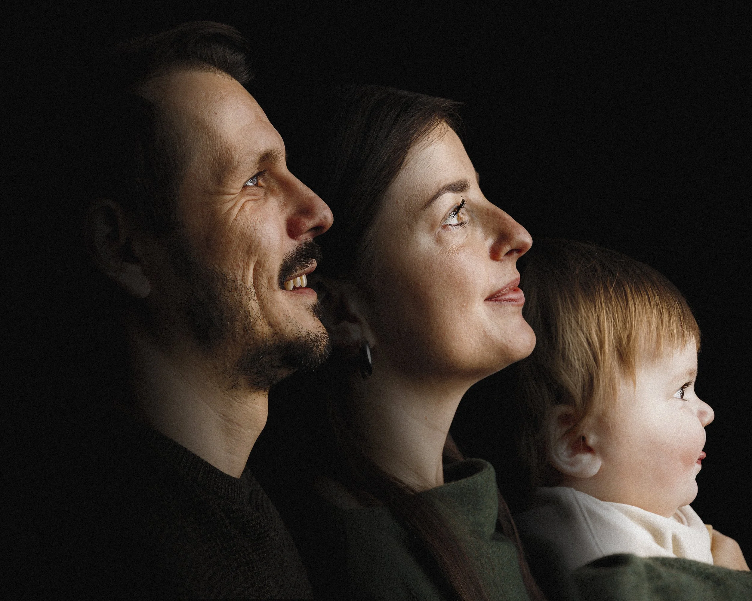 Profile of a family of three, including a man, woman, and small child, all looking to the right against a black background.
