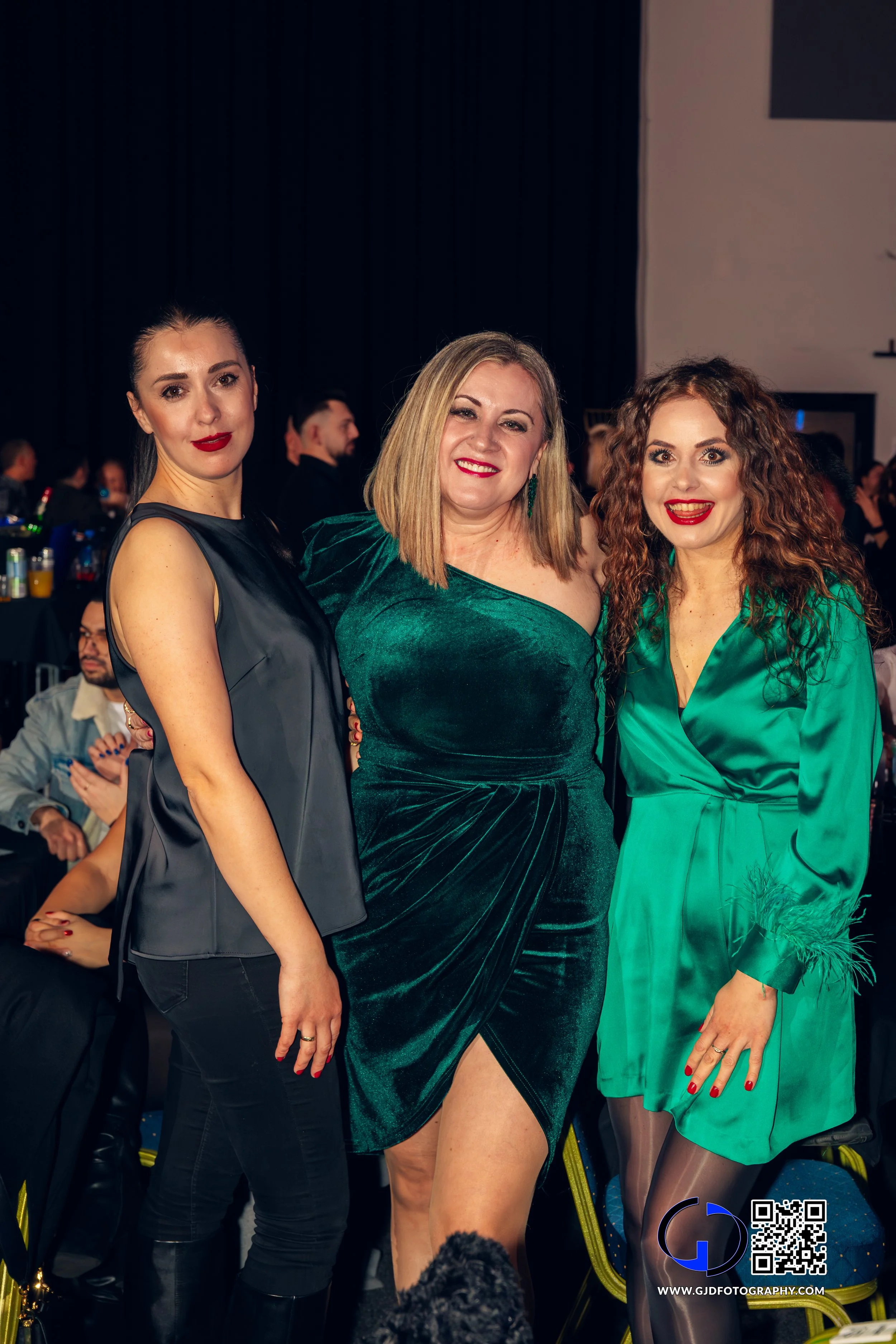 Three women dressed elegantly at a formal event, posing together and smiling for the camera.