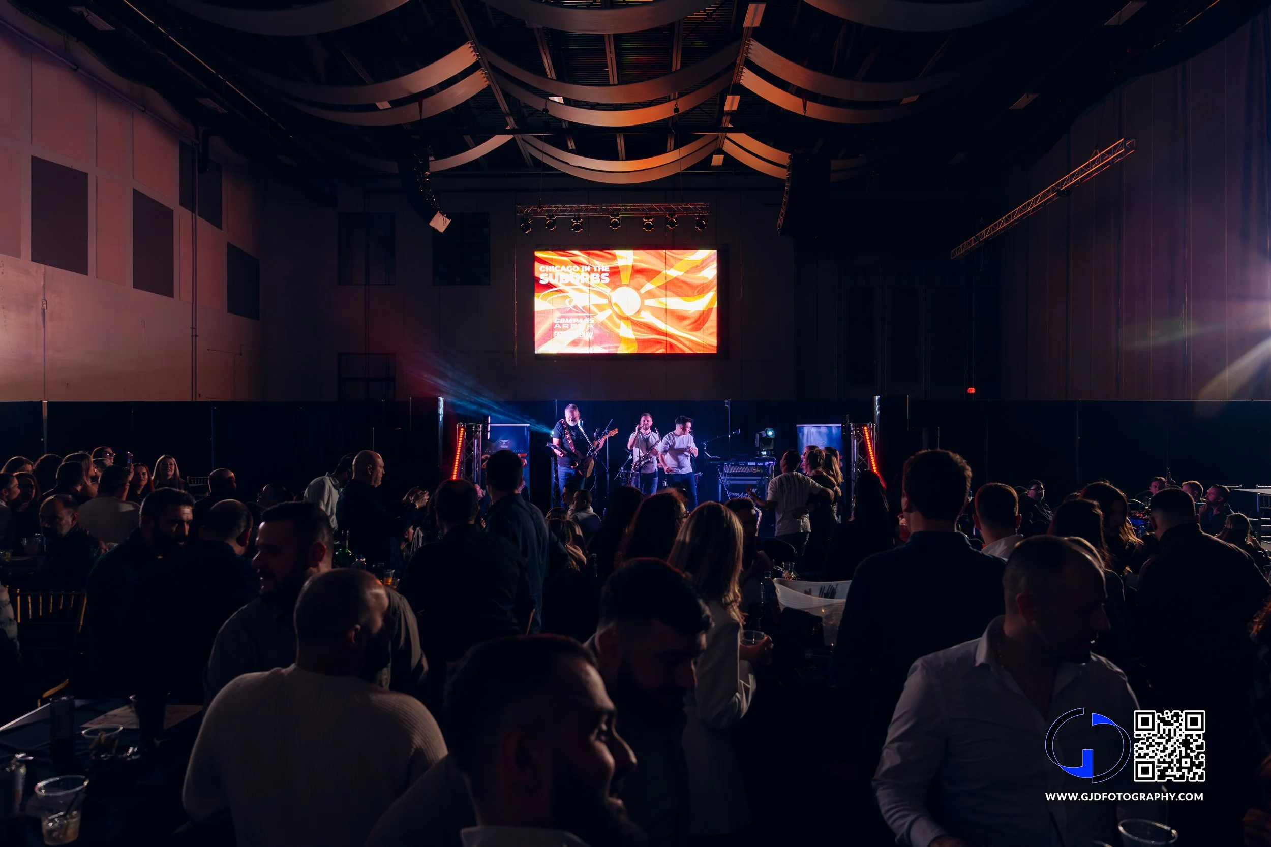 A live band performs on stage at an indoor concert with an audience seated at tables, and a large screen displaying an animated flag behind them.
