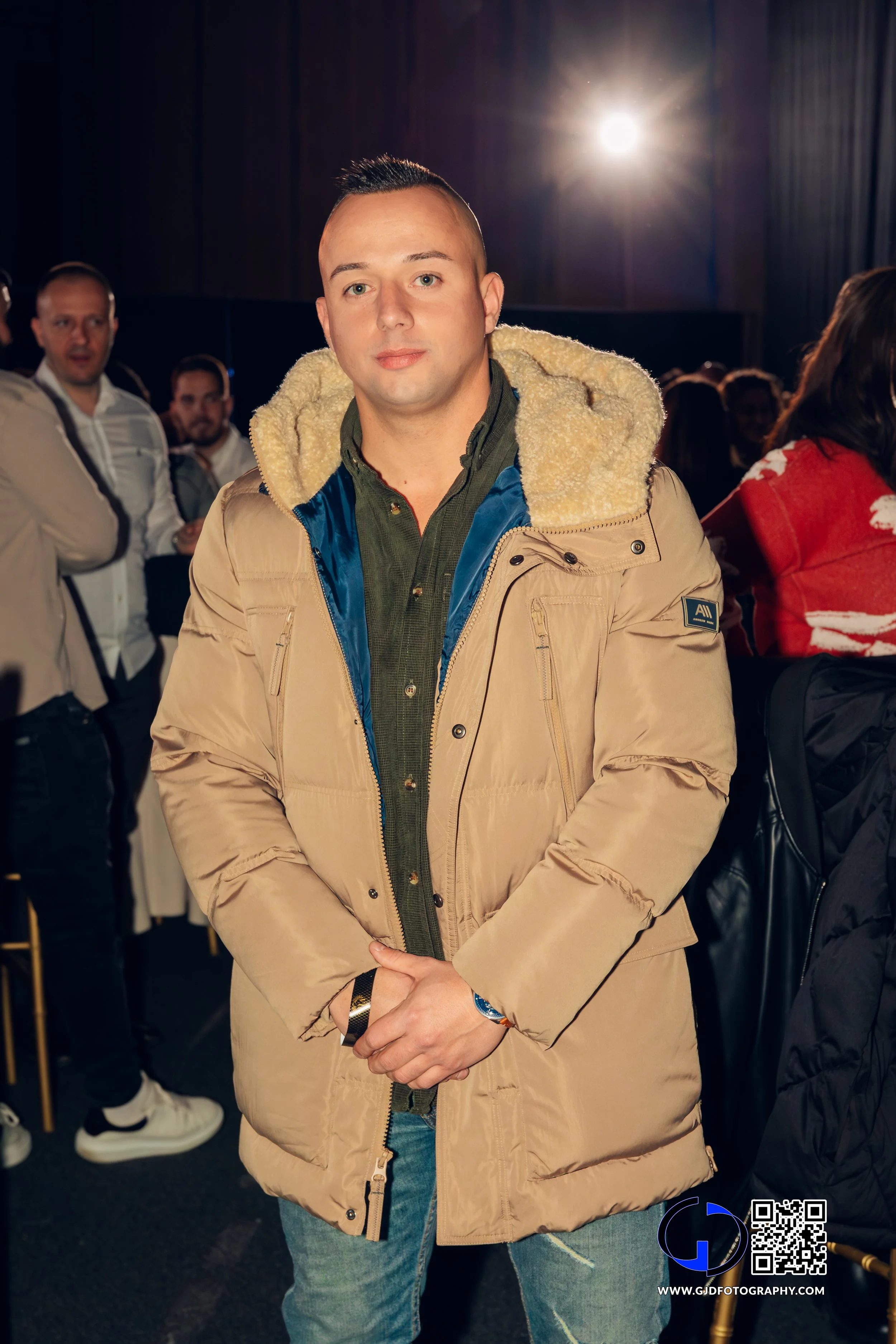 A young man in a beige winter jacket with a fleece-lined hood, standing indoors at a crowded event, with a bright light in the background.