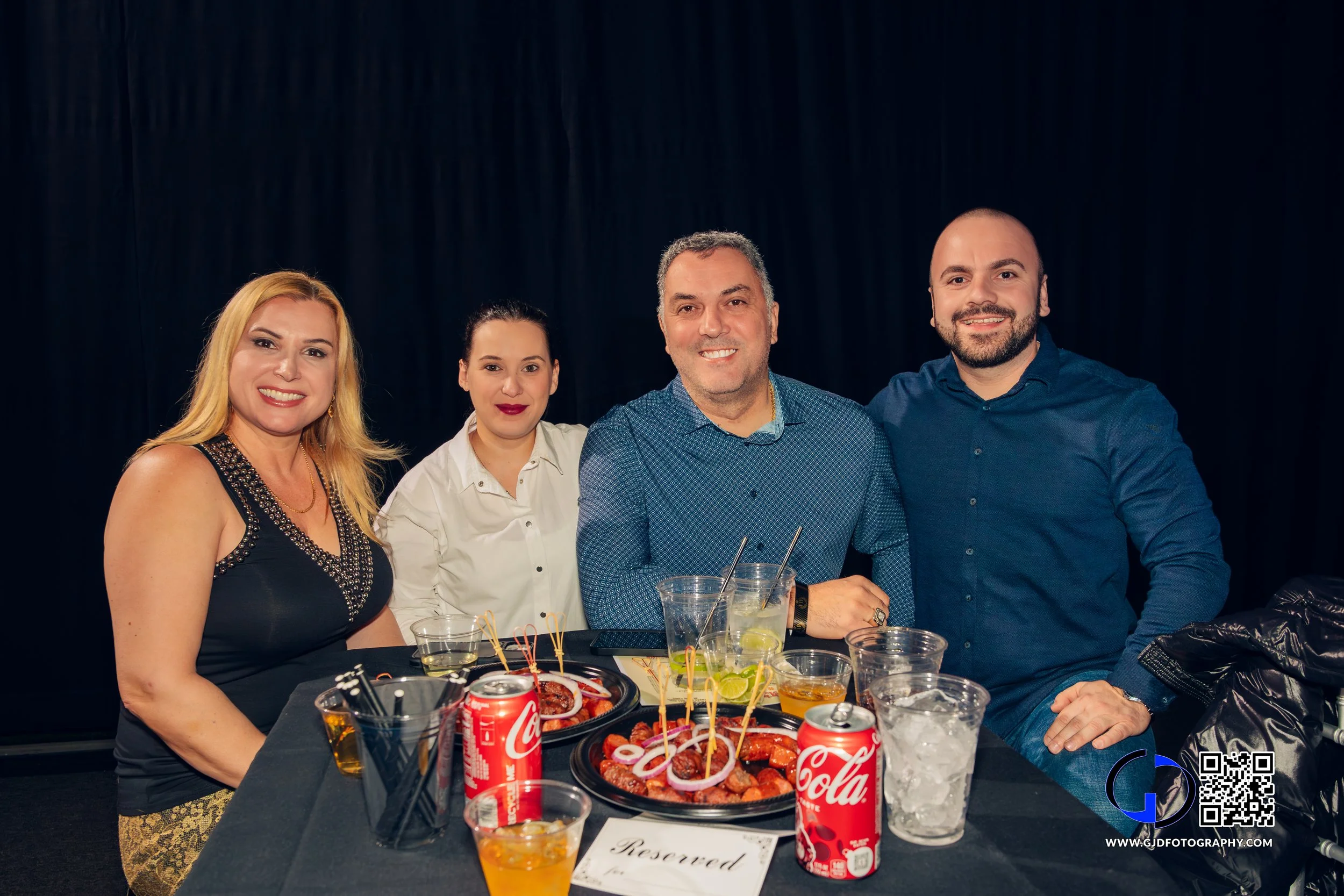 Four people sitting at a table with food and drinks, smiling for the camera at an indoor event.