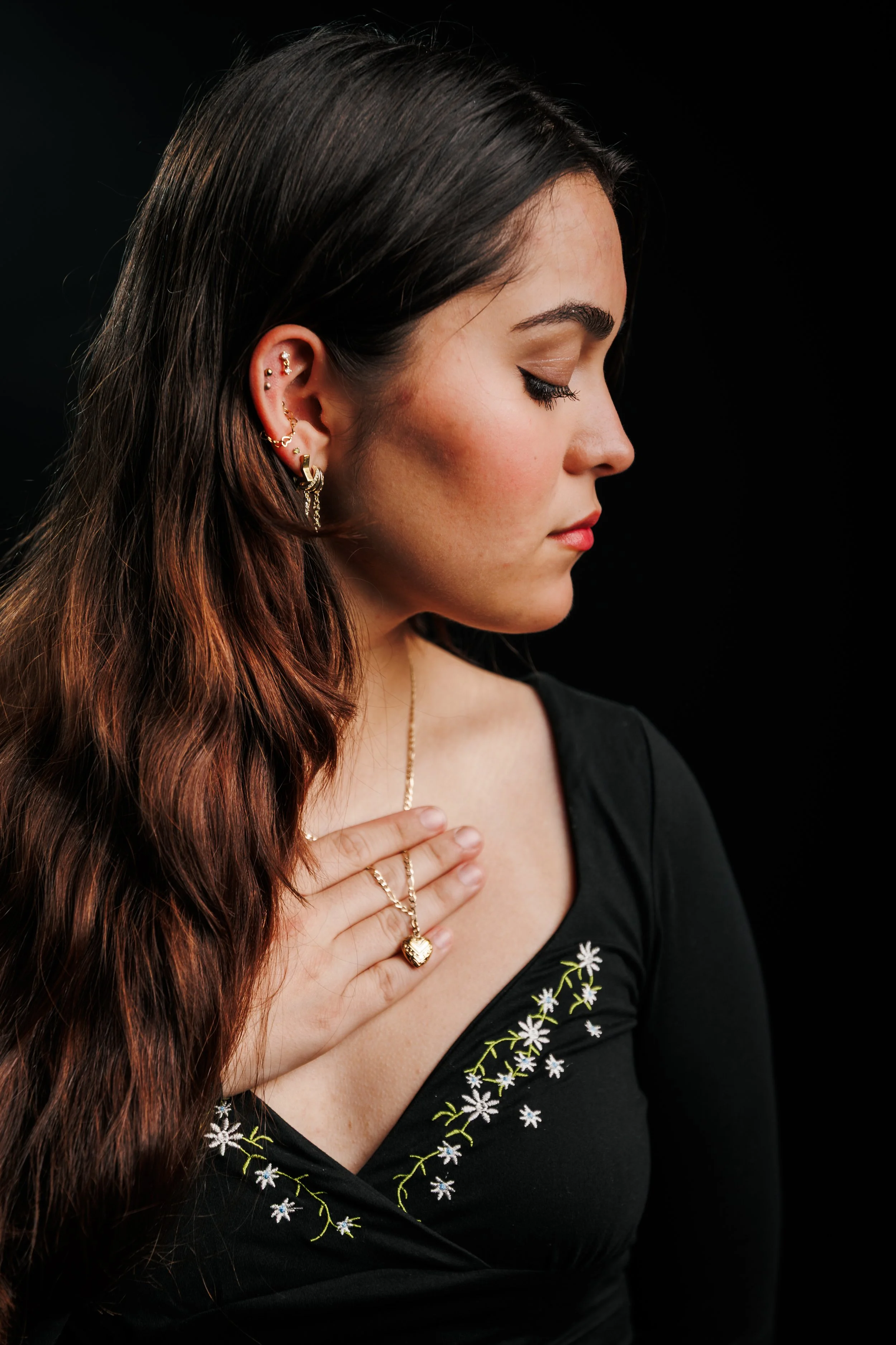 A woman with long, brown hair and multiple earrings, wearing a black top with floral embroidery, and holding a gold heart-shaped pendant necklace near her chest with her right hand, against a dark background.