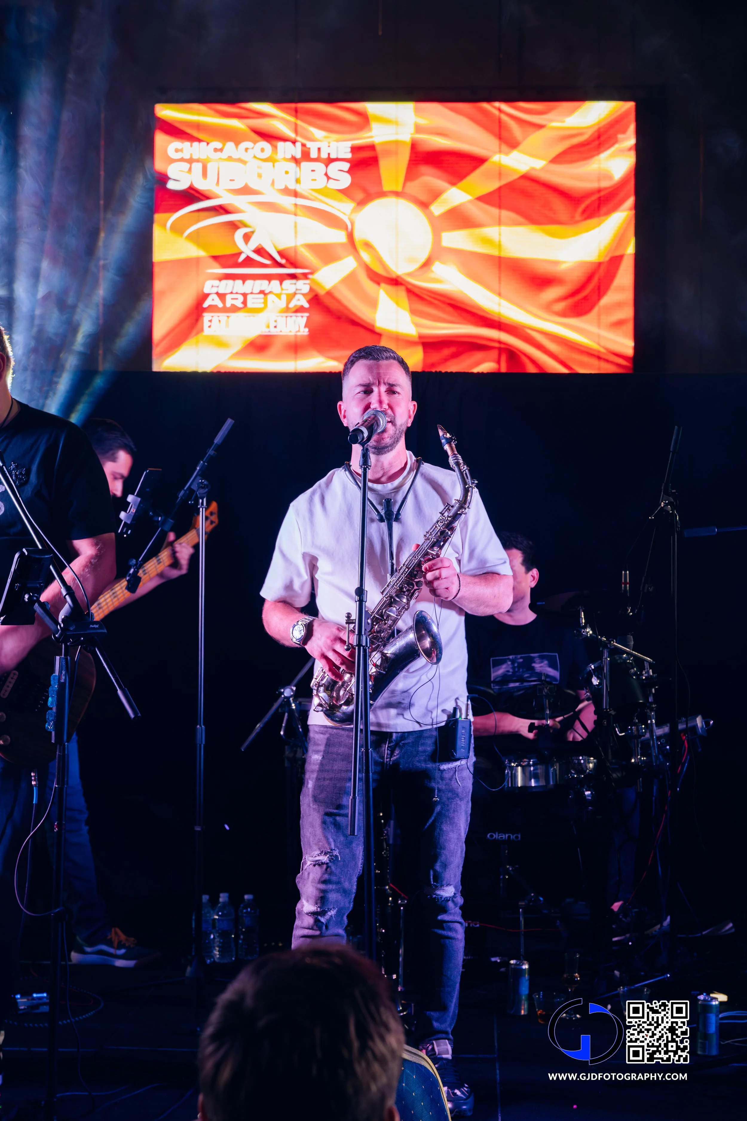 Musician playing saxophone on stage during a live performance with a screen in the background displaying 'Chicago in the Suburbs', 'Compass Arena', and a stylized sun logo.