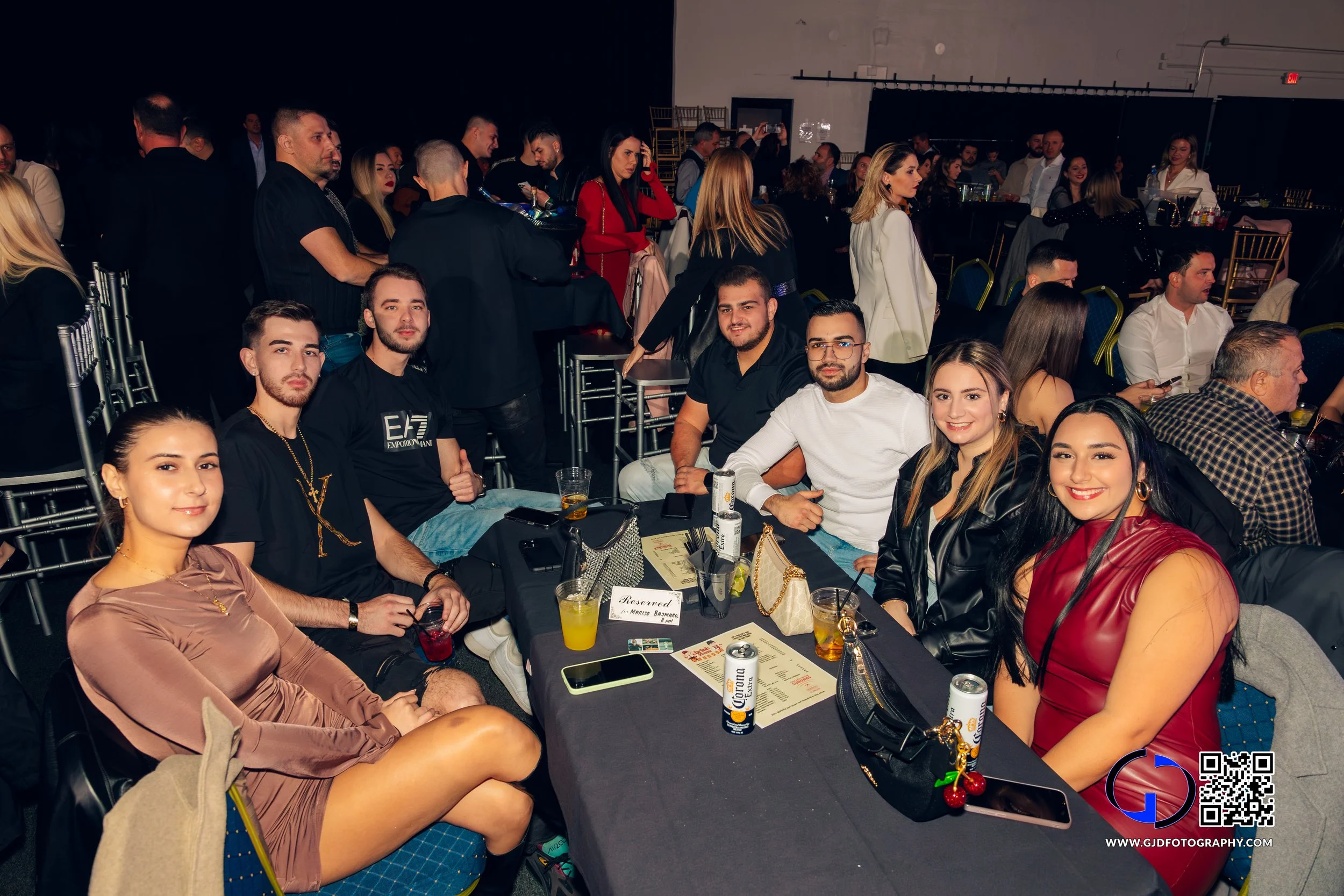 Group of seven young adults sitting around a table at an event, with other people mingling in the background.