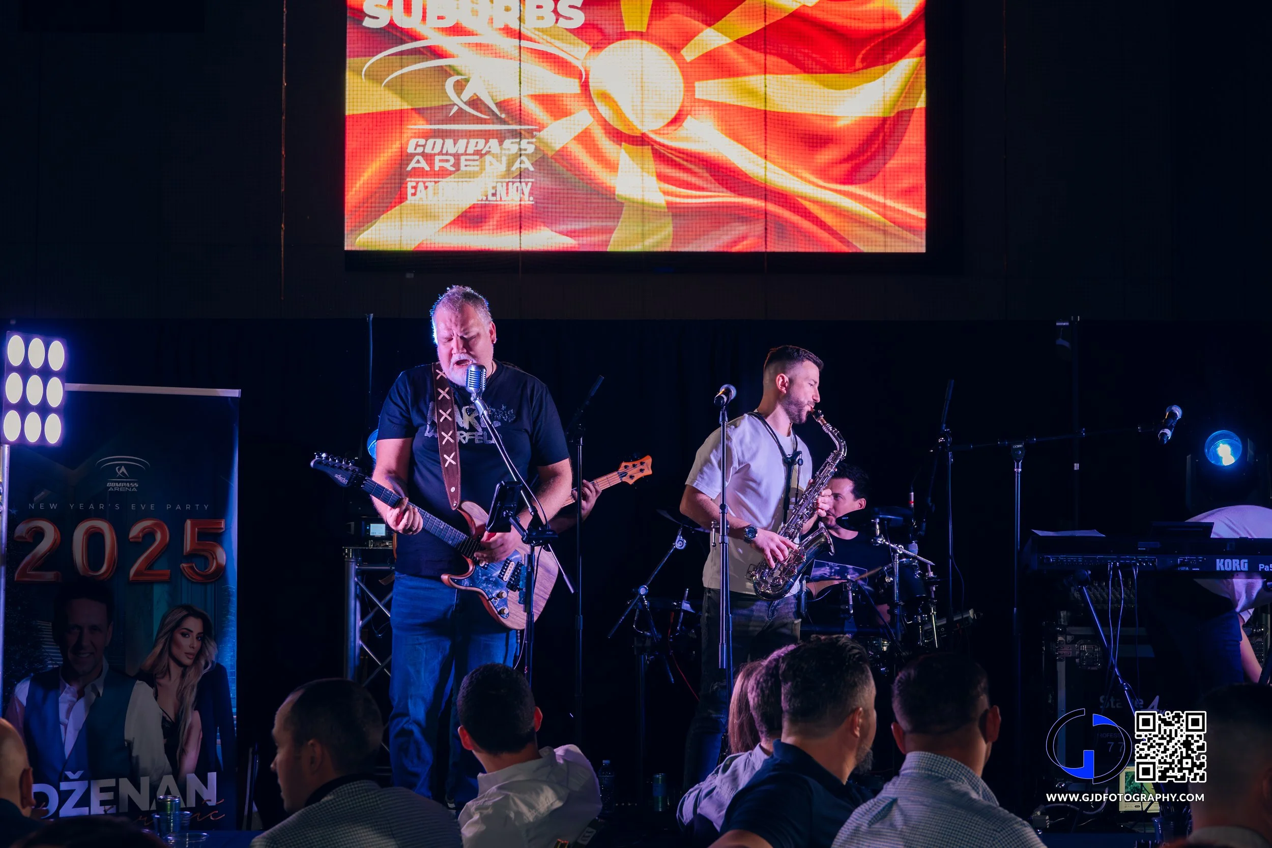 Musicians performing on stage at a New Year's Eve party, playing guitar, saxophone, and drums, with a large screen displaying graphics and a poster on the side promoting the event and new year 2025.