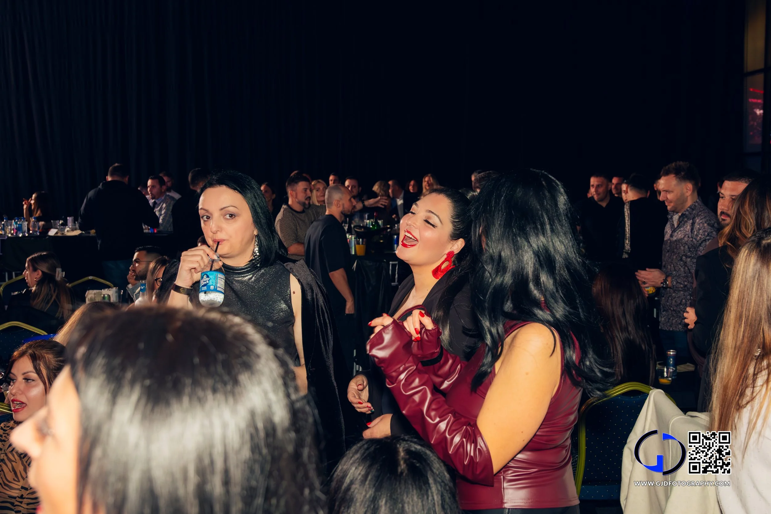 Group of people at a social event, some women in the foreground smiling and holding drinks, men in the background socializing, indoor venue with dark curtains