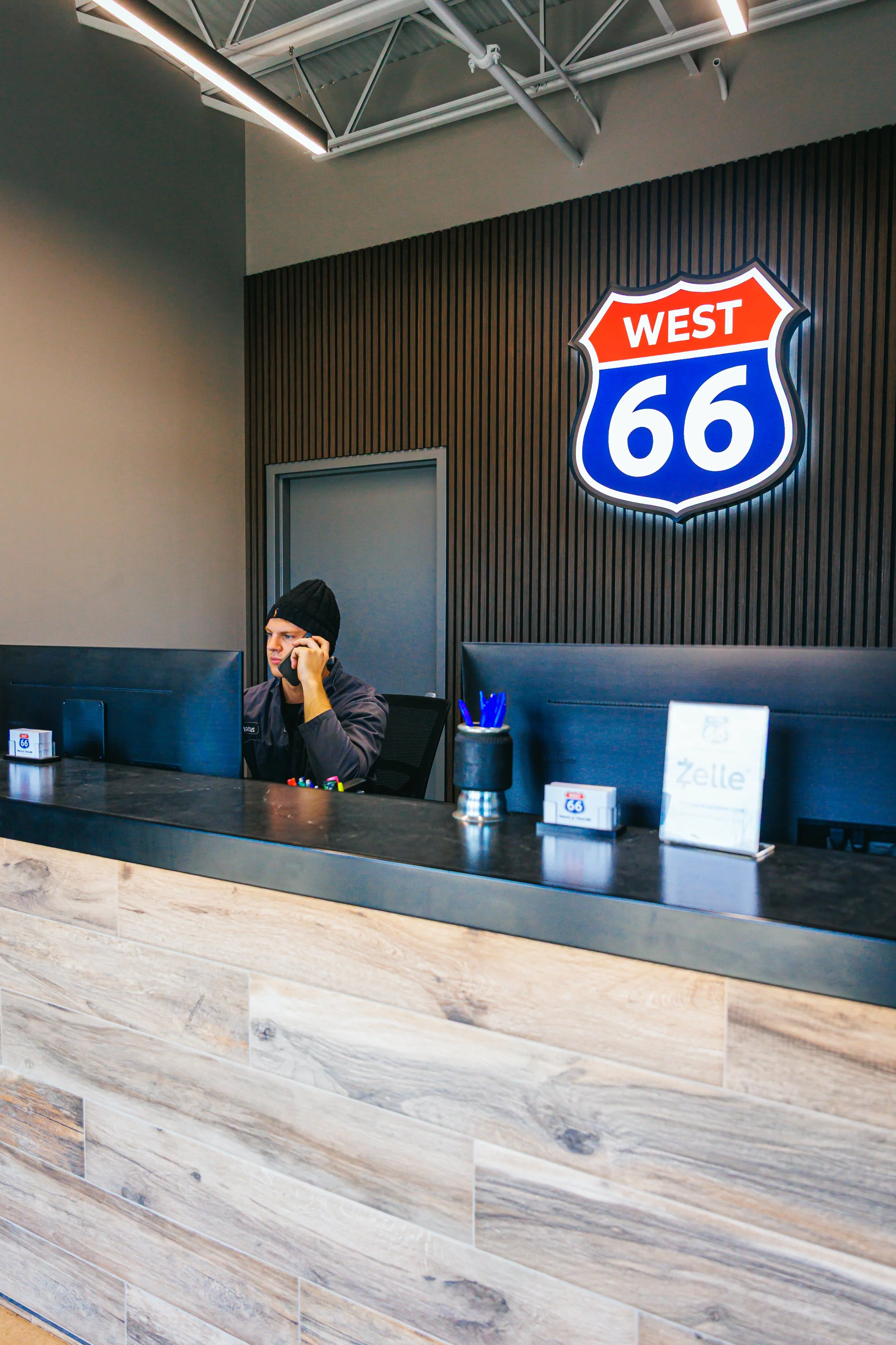 Reception desk with a young man on the phone, illuminated 'West 66' highway sign on the wall behind.
