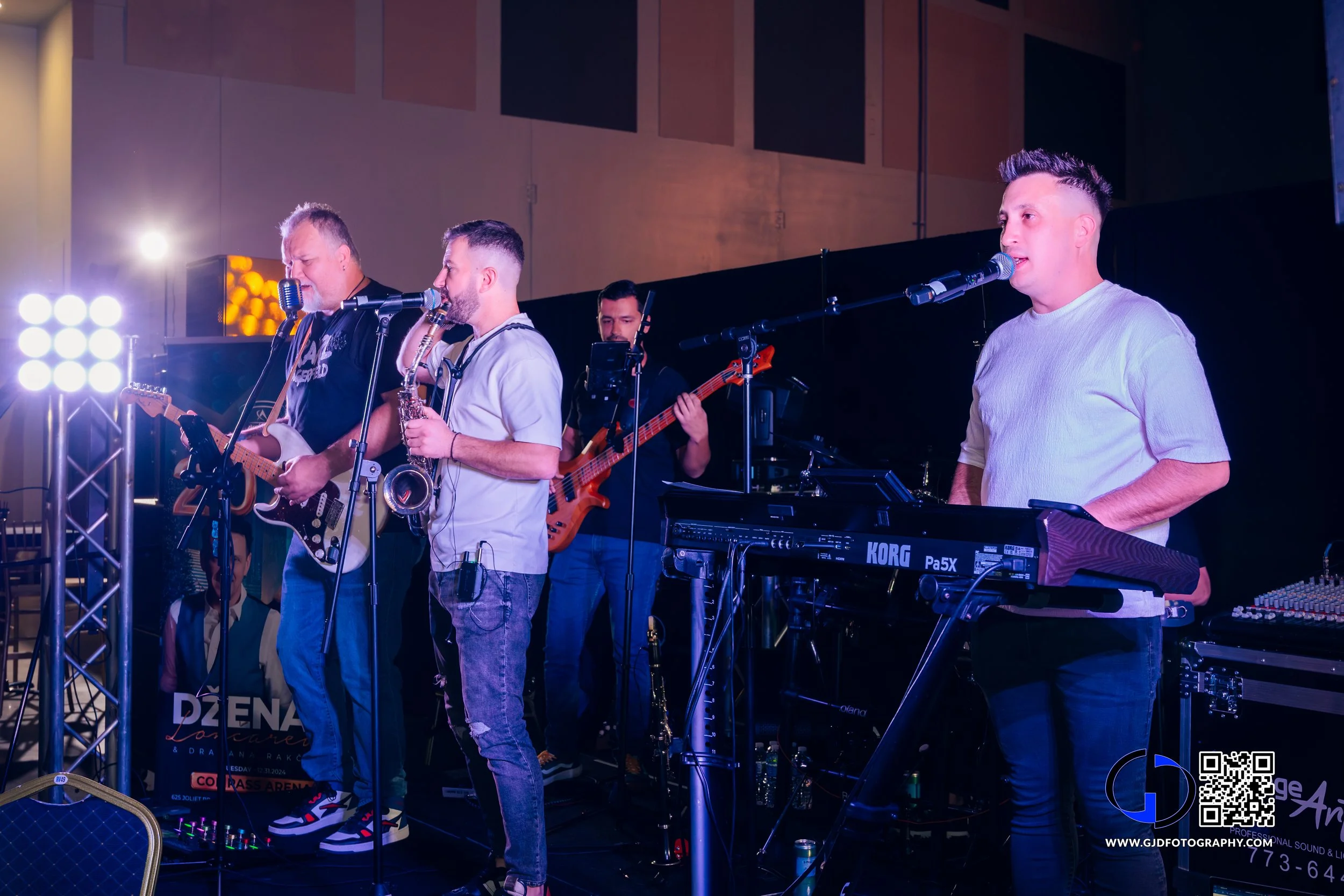 A band performing on stage with four musicians, including a guitarist, a saxophonist, a bassist, and a keyboard player, at an indoor event.