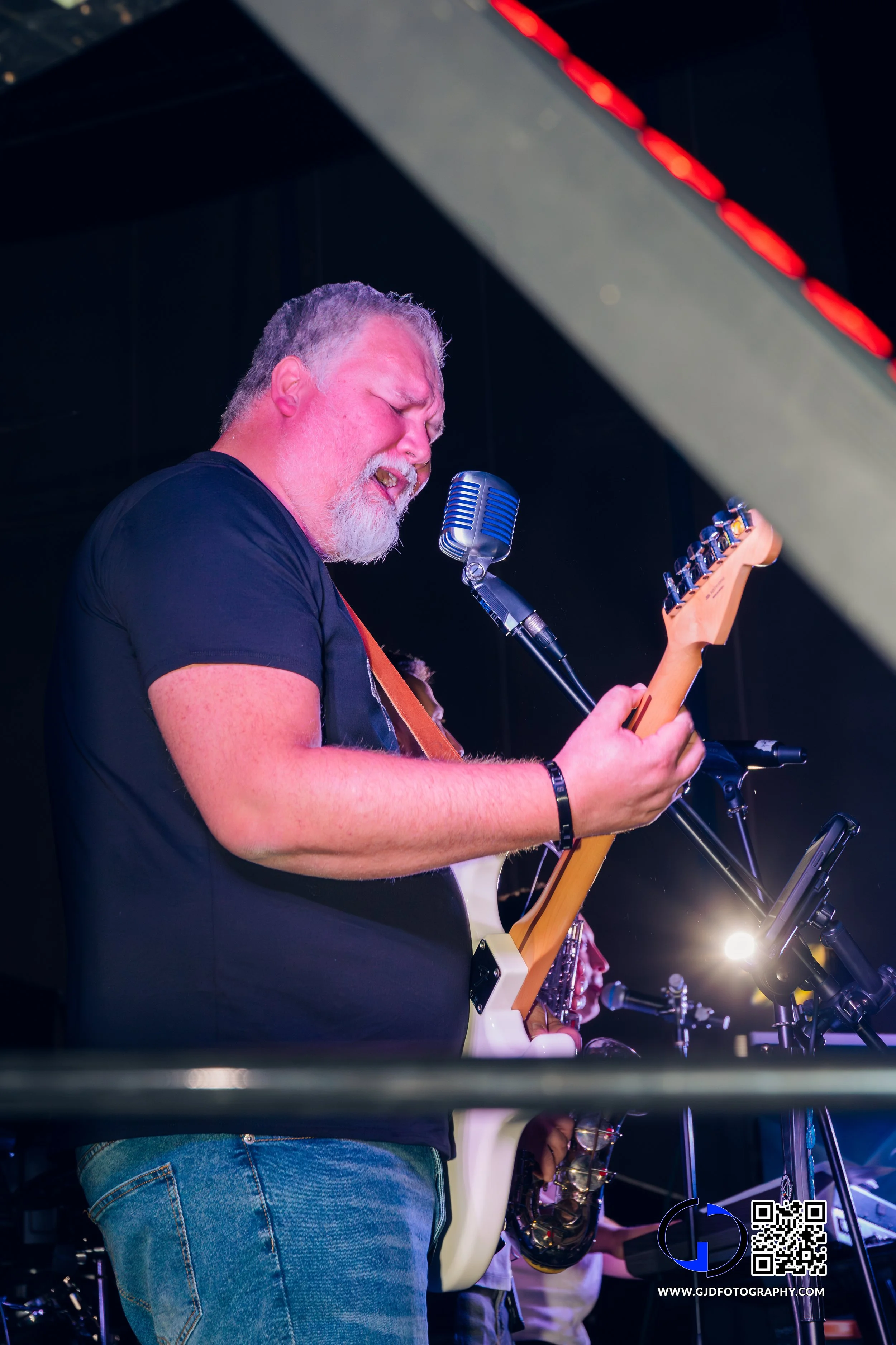 A man with a beard and gray hair wearing a black t-shirt playing an electric guitar with a vintage style microphone in front of him on stage, with a saxophonist in the background.