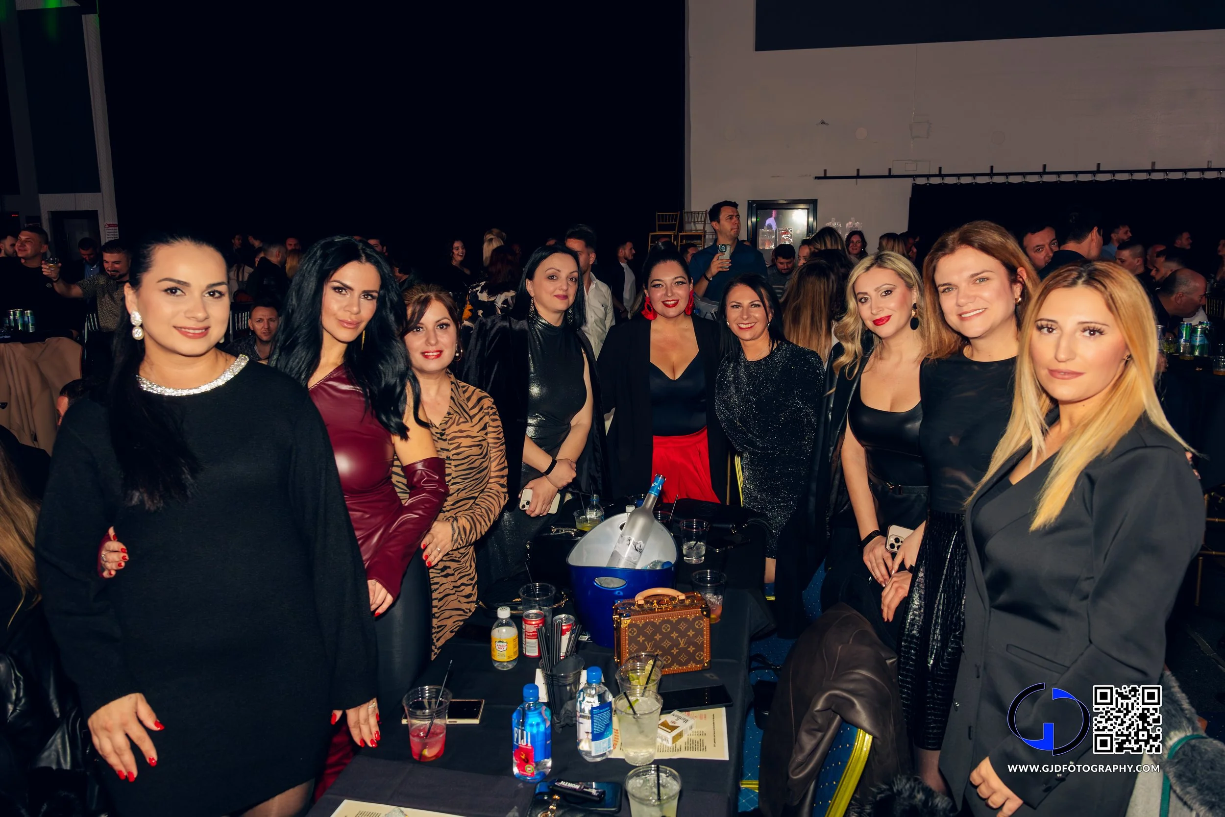 Group of women dressed in elegant clothing at a social event or party, standing around a table with drinks and a blue ice bucket, in a large indoor venue with many other attendees in the background.