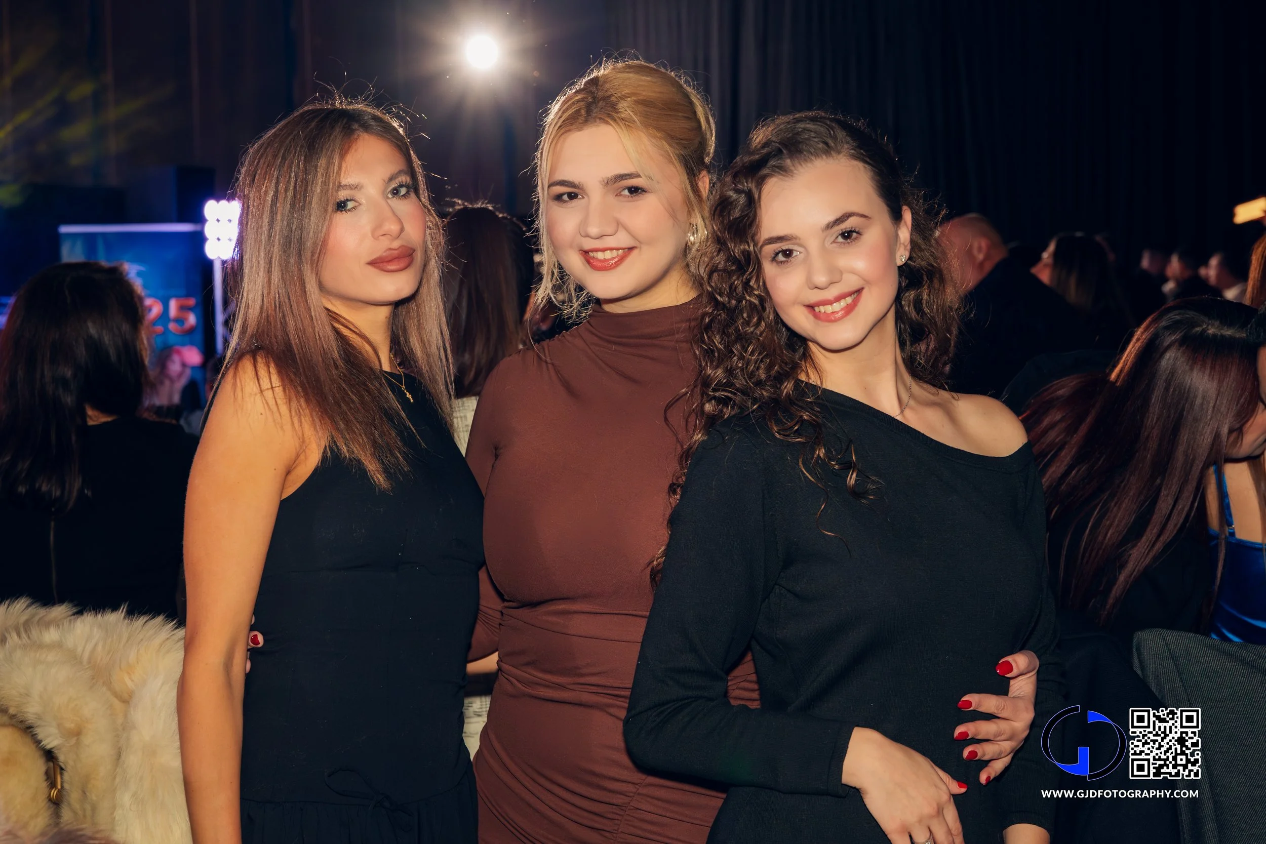 Three young women smiling and posing for a photo at an indoor event with a crowd in the background.