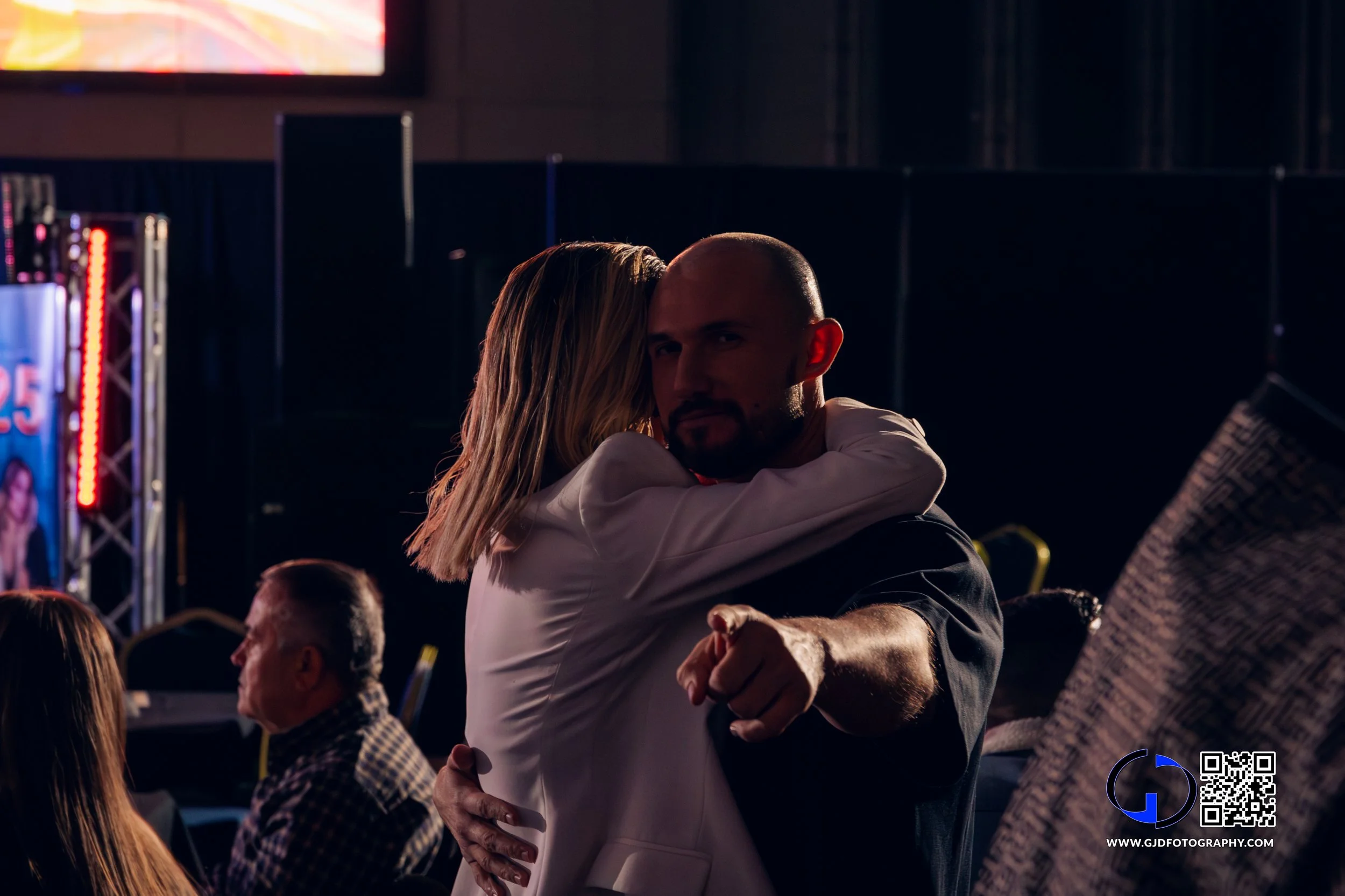 A man and woman hugging, with the woman wrapping her arms around the man's neck at a dimly lit event or party.