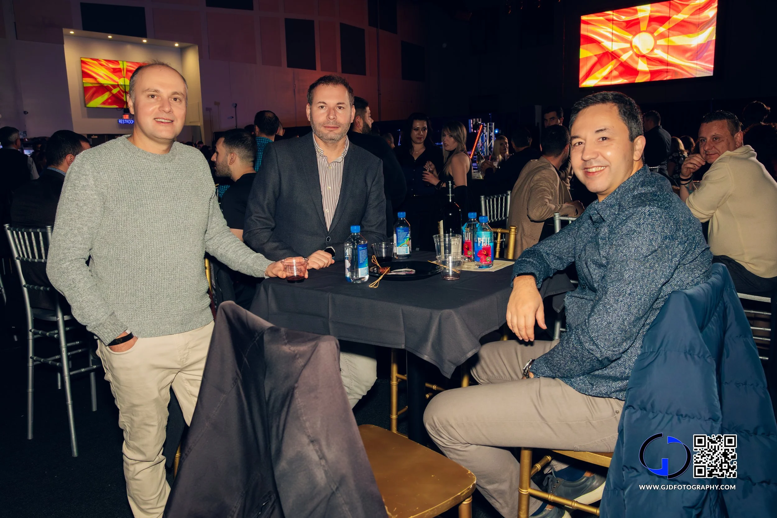 Three men at a round table with drinks, in a large, crowded event hall with a large screen displaying a red and yellow design in the background.