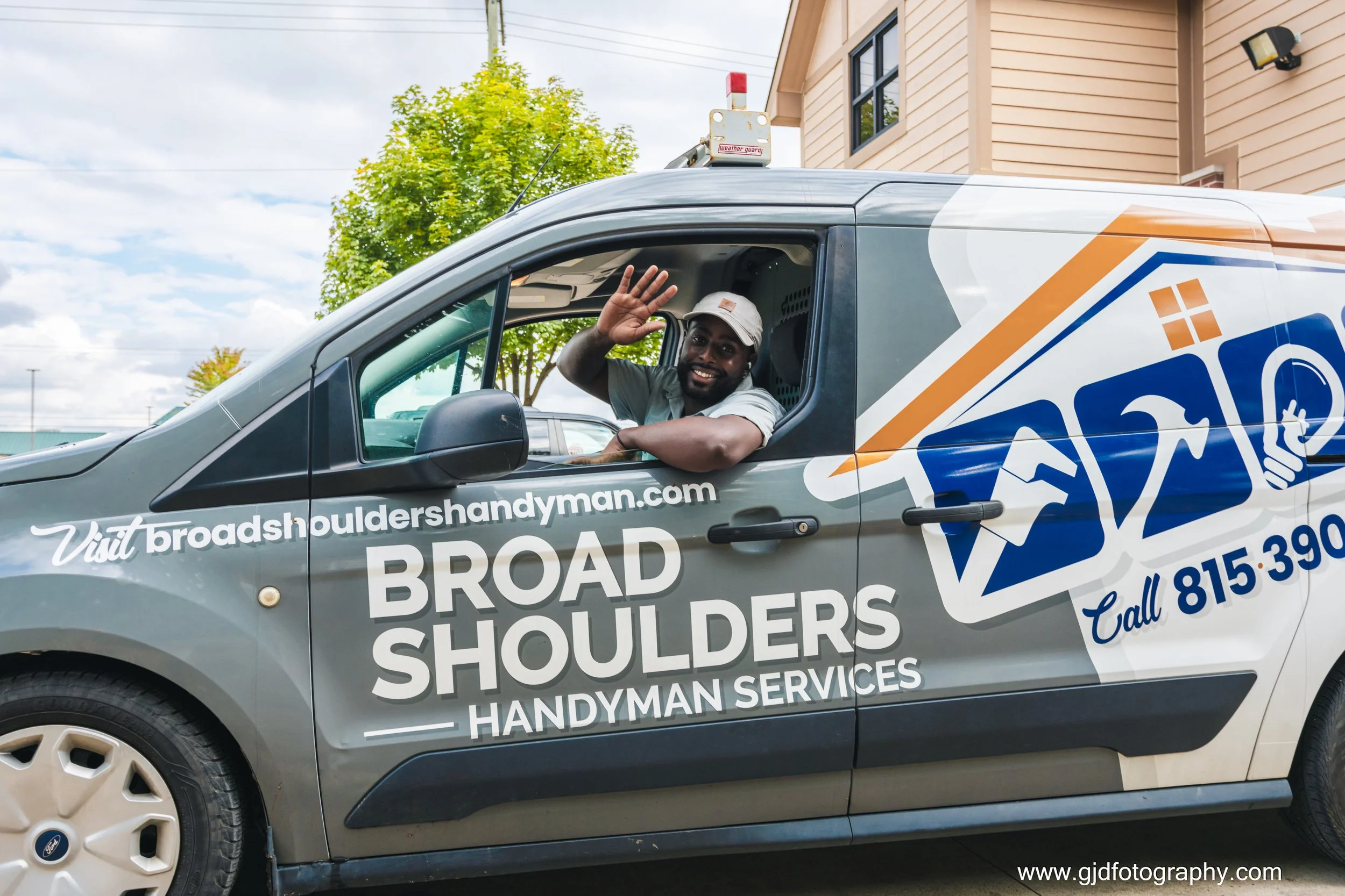 A smiling man waving from the driver's window of a company van labeled 'Broad Shoulders Handyman Services' parked outside a residential building with trees in the background.