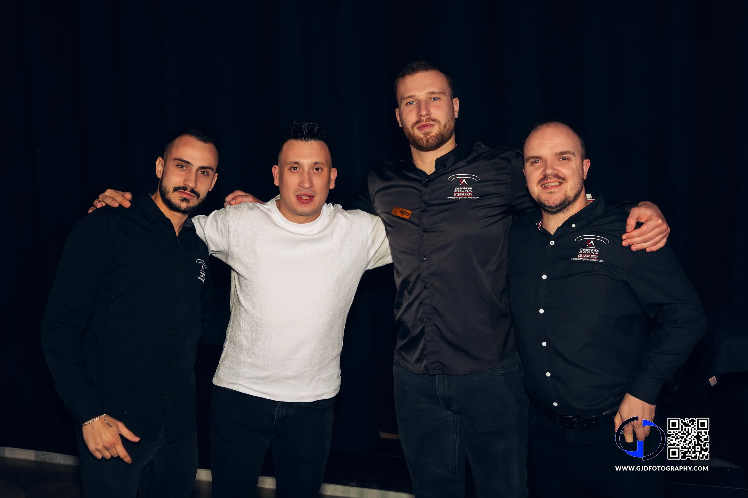 Four men standing together in front of a black background, with the two on the ends wearing black shirts and the two in the middle wearing white and black shirts, smiling for the camera.