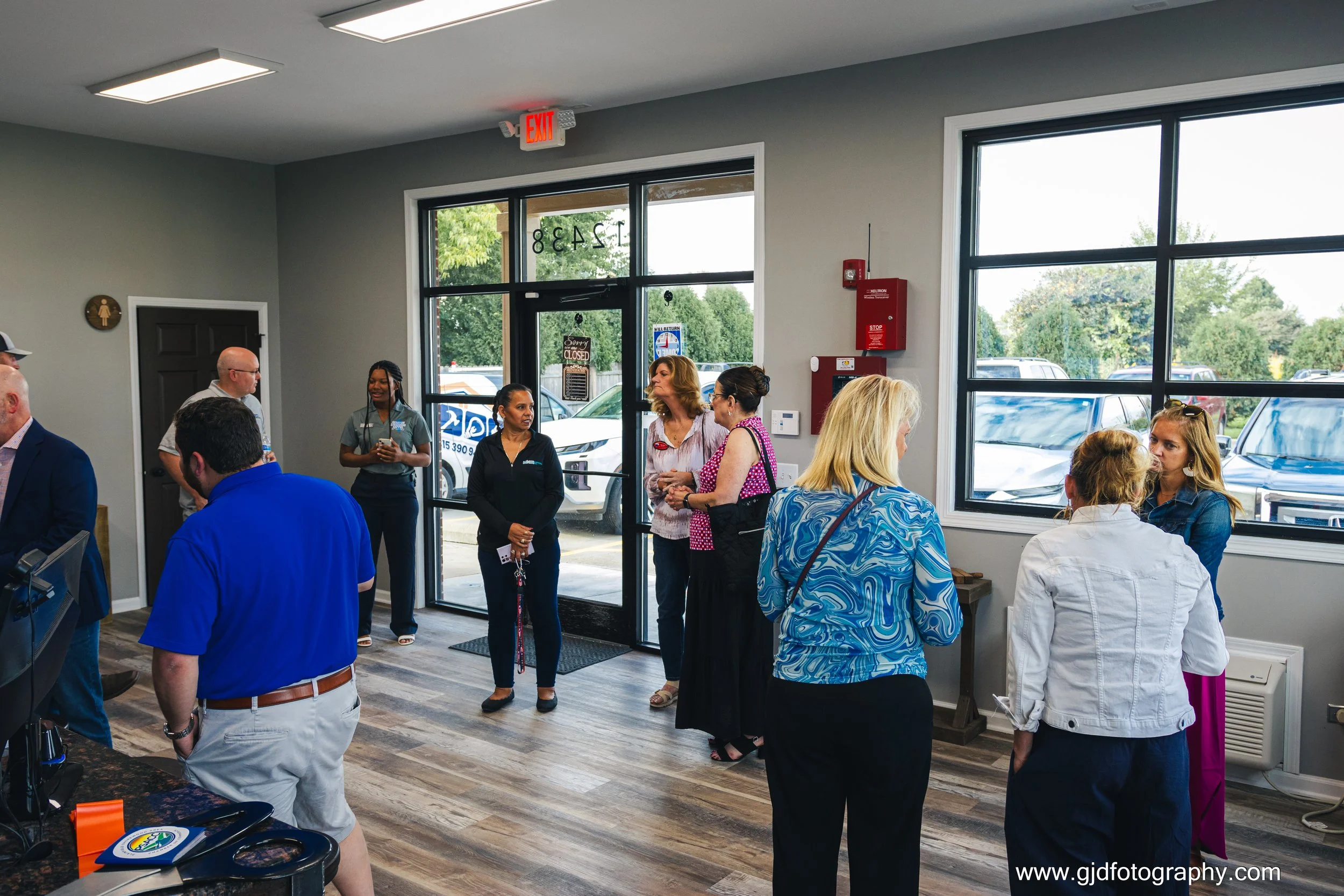 People standing inside a building near a large window, engaged in conversation and waiting. Entry door visible with a car outside.