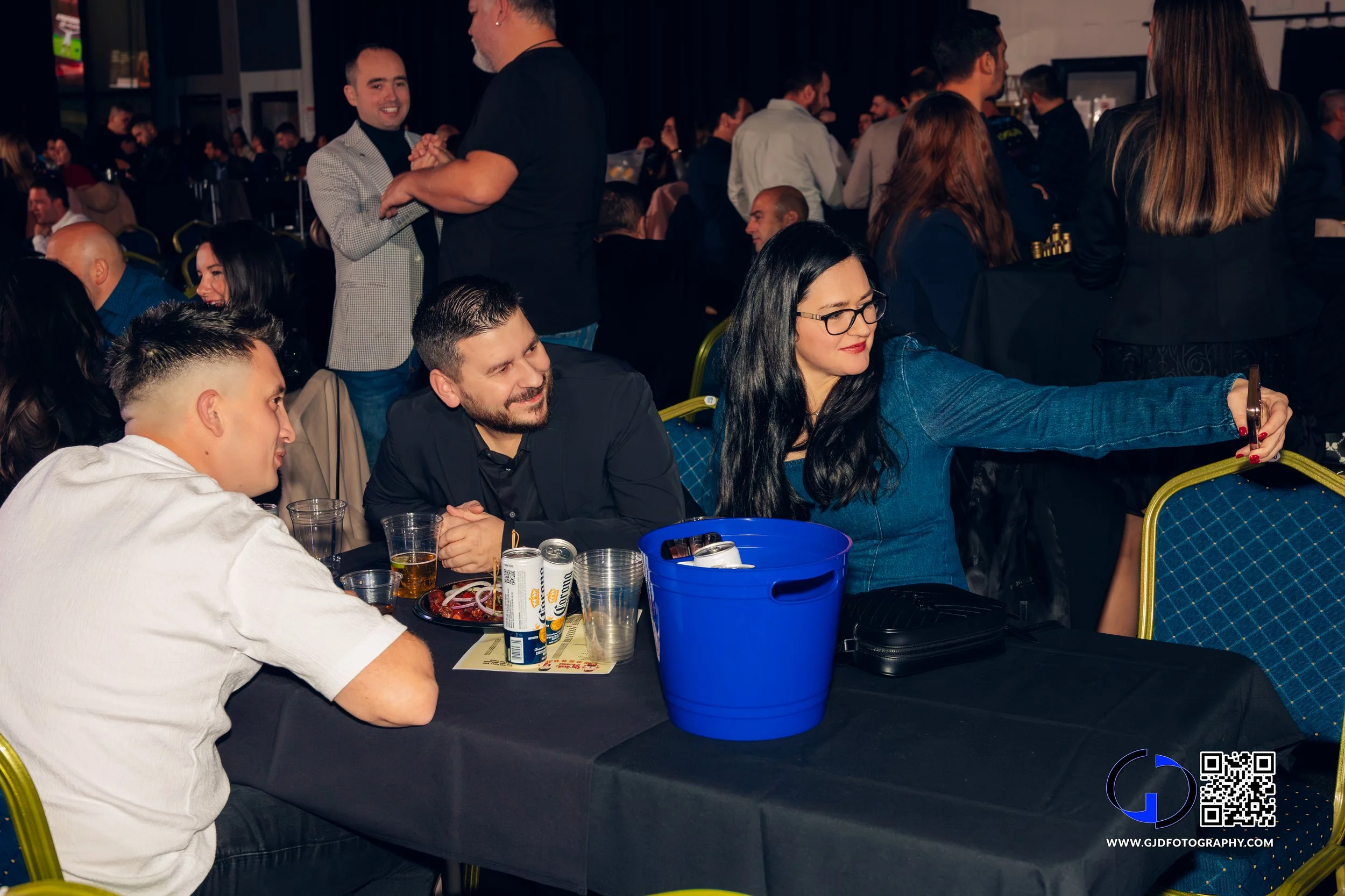 Group of people sitting at a table in a crowded event venue, with some taking a selfie and others talking or smiling.