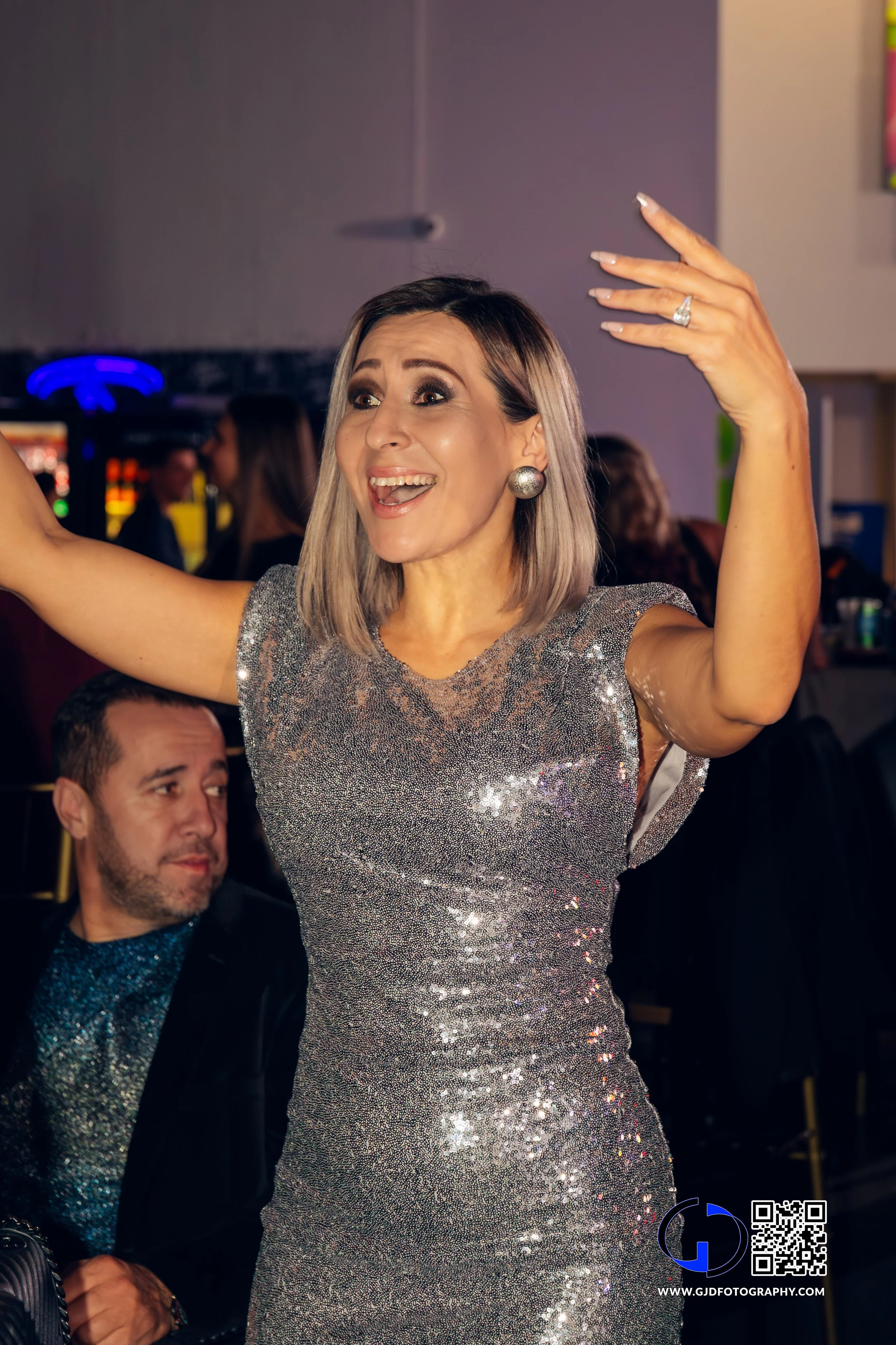 A woman in a sparkly silver dress gestures excitedly and smiles at a party or celebration, with a man sitting nearby in the background.