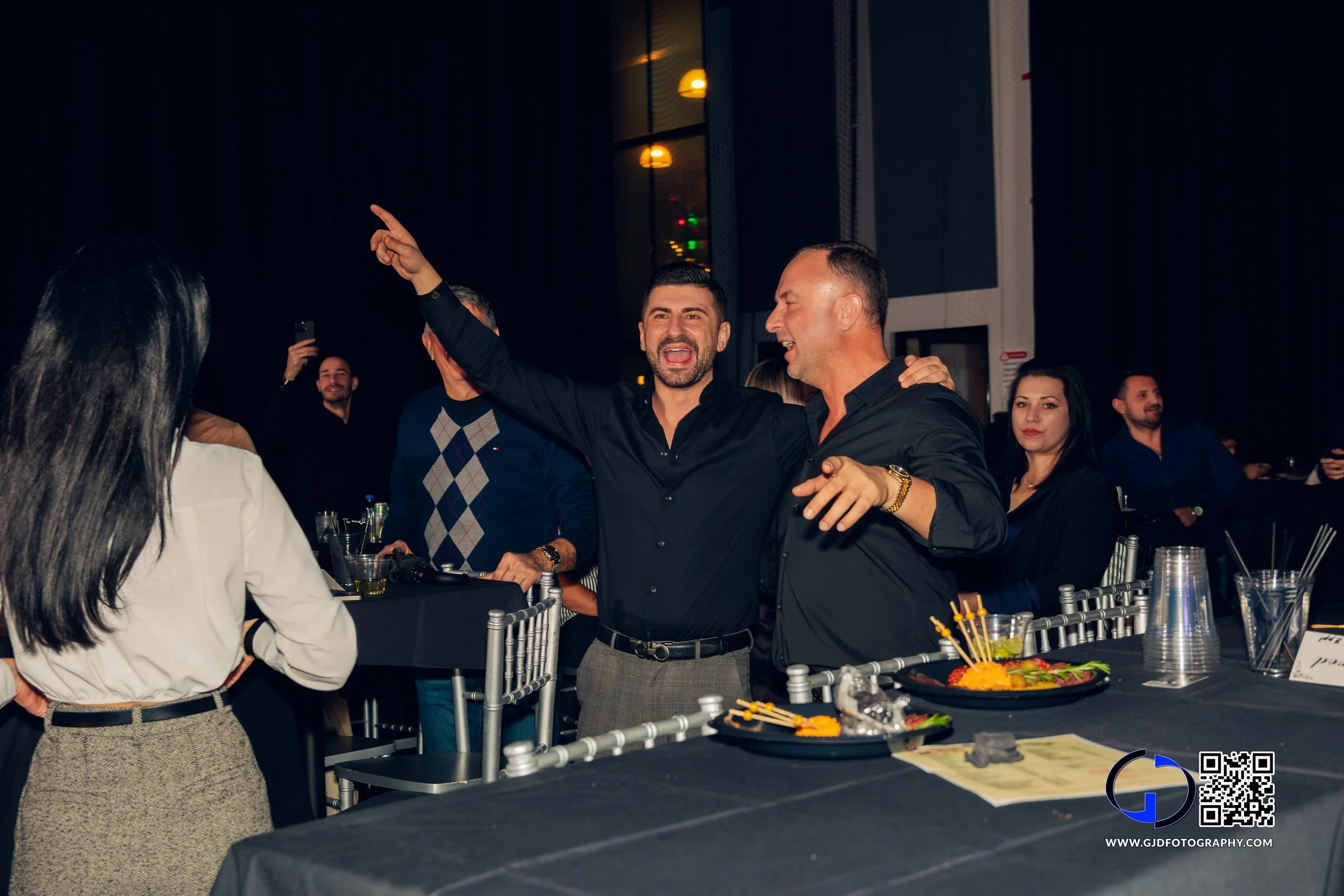 Two men are celebrating at a party, one is pointing upward and the other has his arm around him, surrounded by other guests in a dark room with colorful lighting.