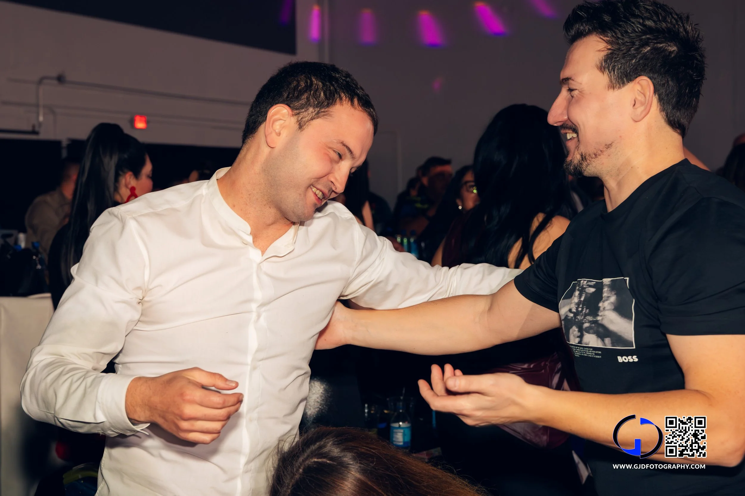 Two men smiling and laughing while dancing at a party or celebration. One wears a white shirt, and the other wears a black T-shirt with a graphic print. They are surrounded by other people in a dimly lit room with purple lighting.