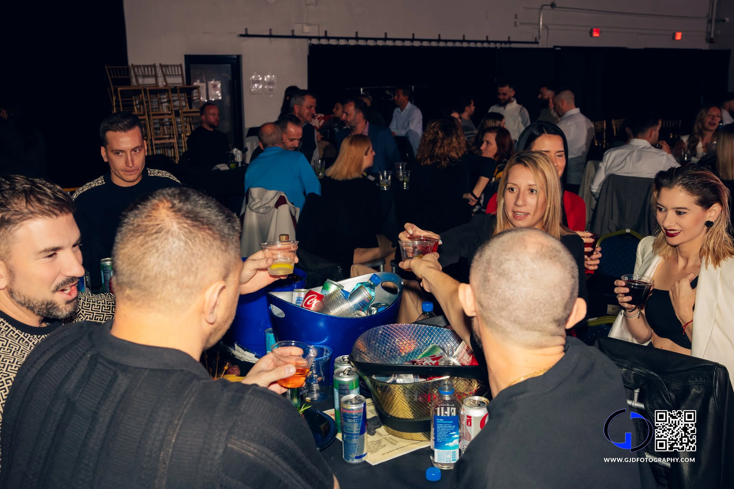 Group of people at a party or social gathering, sitting around a table with drinks, engaging in conversations and smiling.