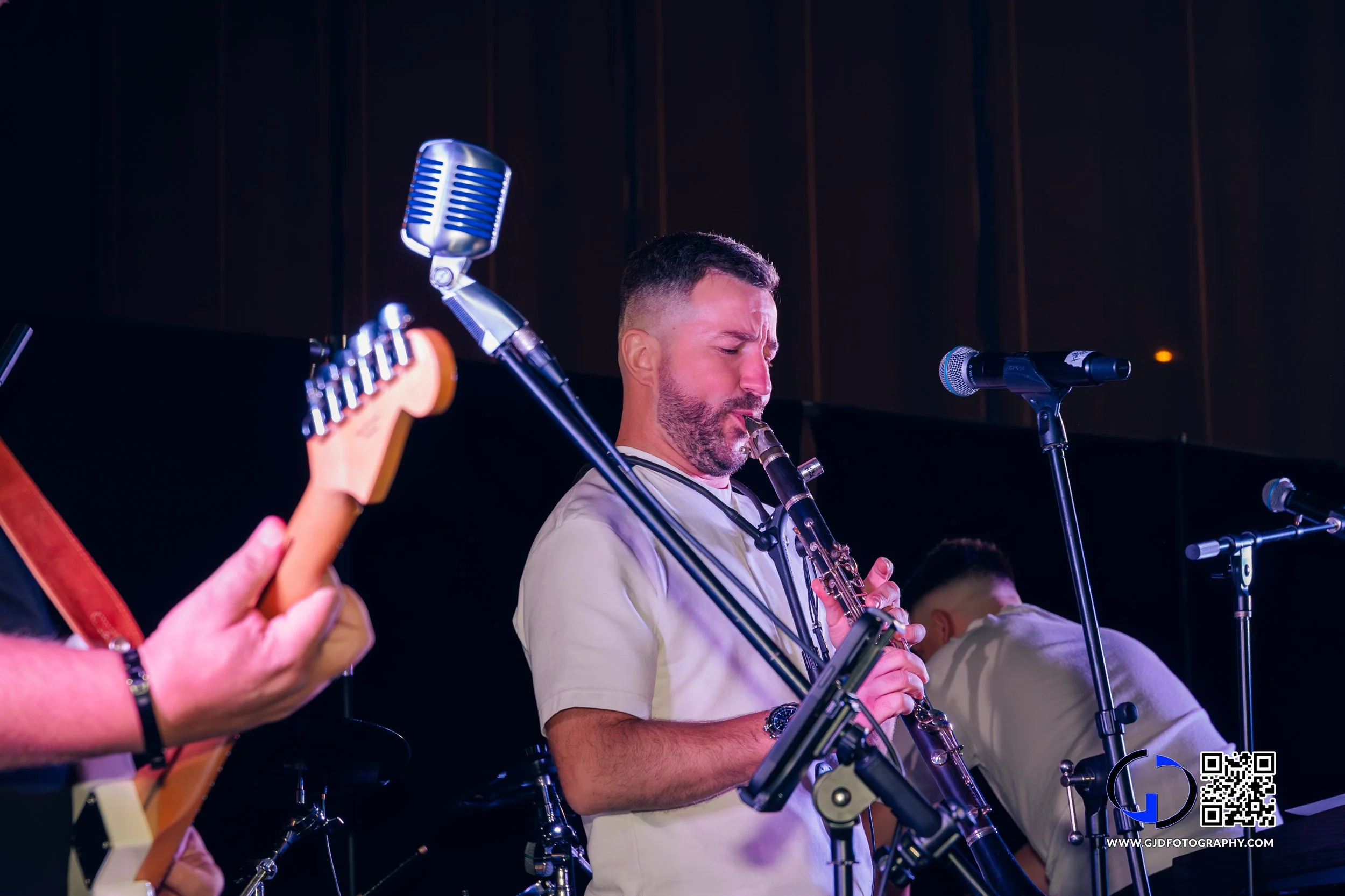 Musician playing the saxophone during a live performance, surrounded by microphones and another musician playing a guitar