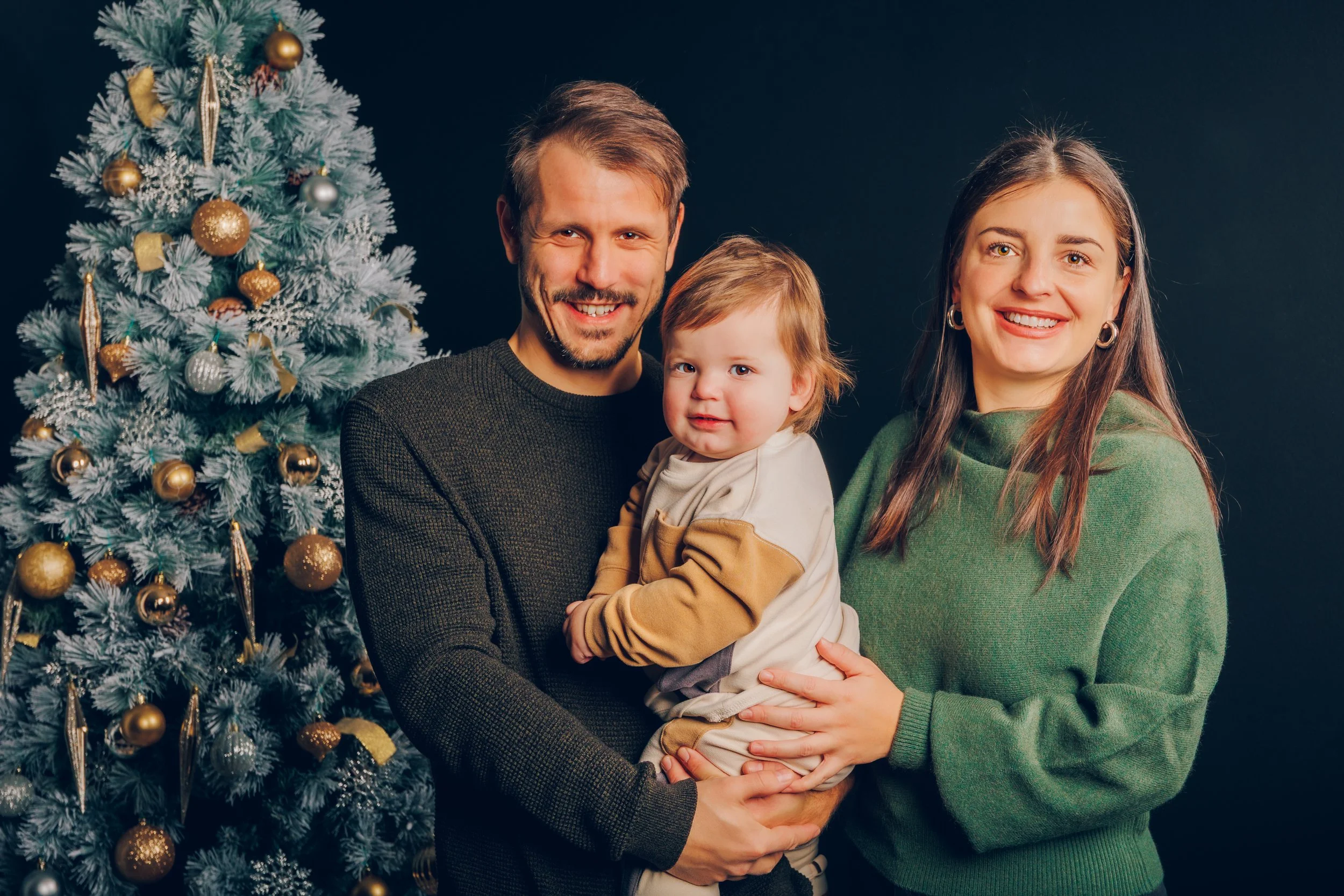 A smiling family of three, with a father, mother, and young child, standing in front of a decorated Christmas tree during the holidays.