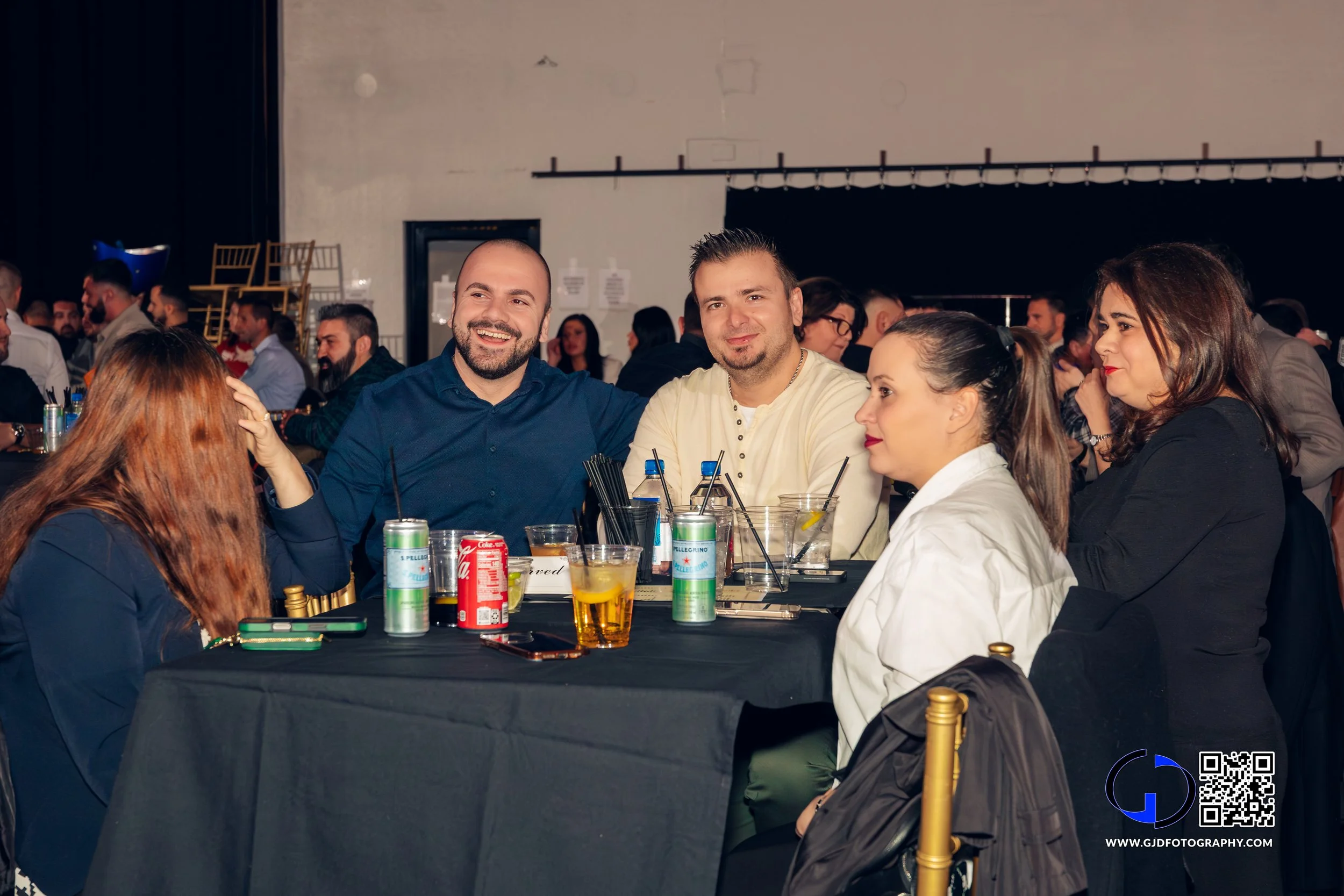 Group of people sitting around a table at a social event, smiling and talking, with drinks and straws on the table, in an indoor venue with a dark backdrop.