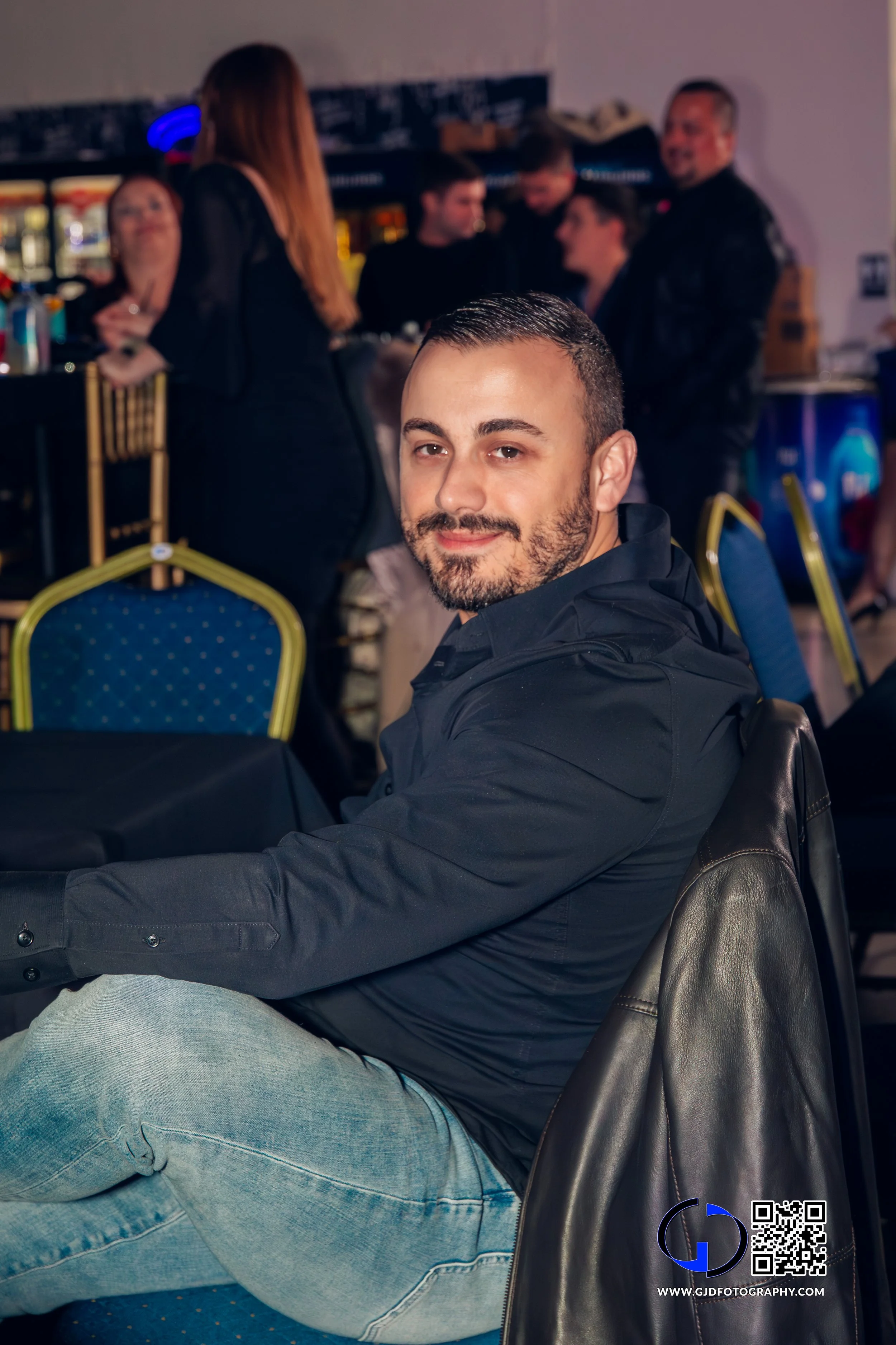 A man with a beard and short hair smiling at the camera, seated at a table in a social setting with a bar and group of people in the background.