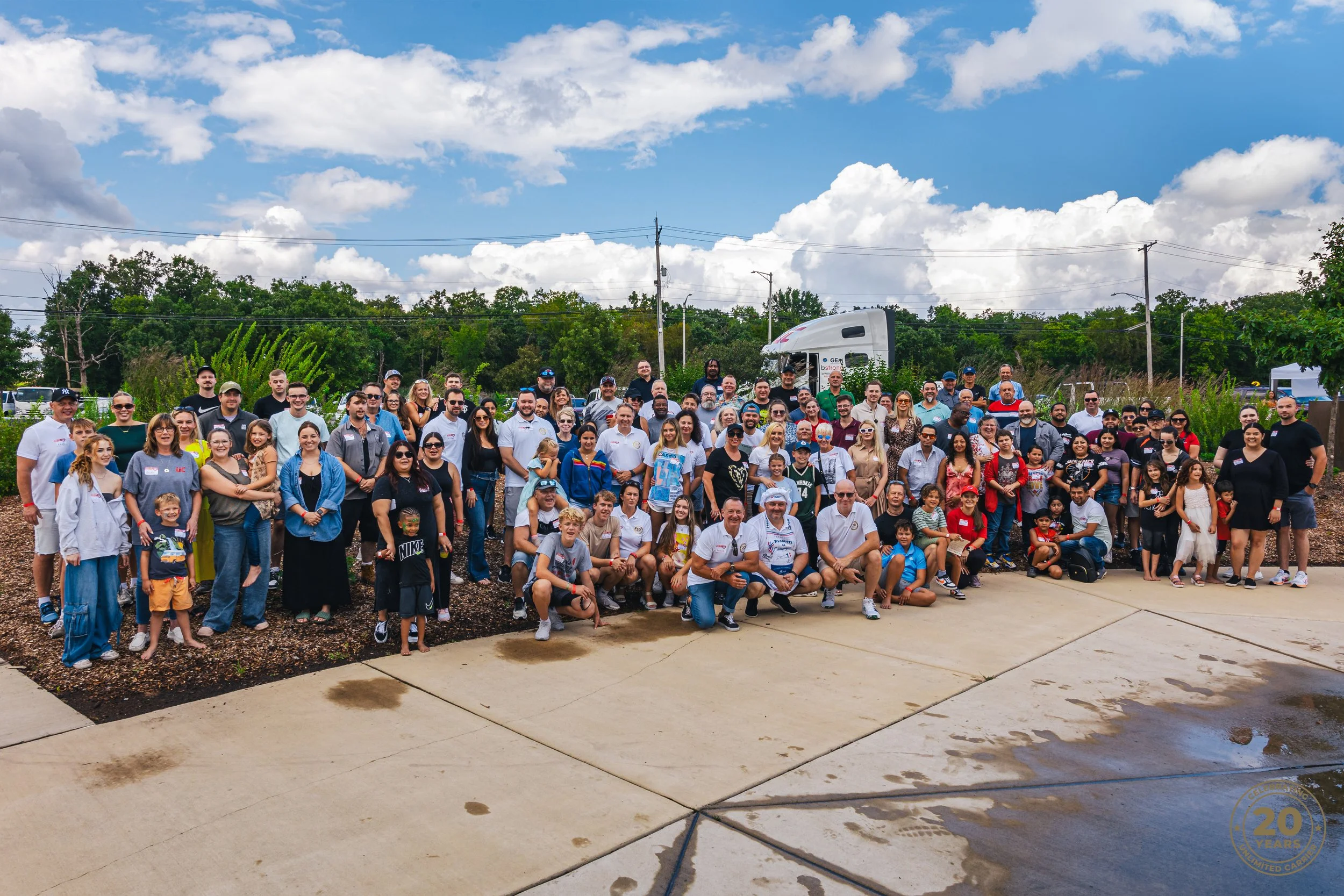 Group of people gathered outdoors on a partly cloudy day, posing for a large photo with trees, power lines, and a truck in the background.