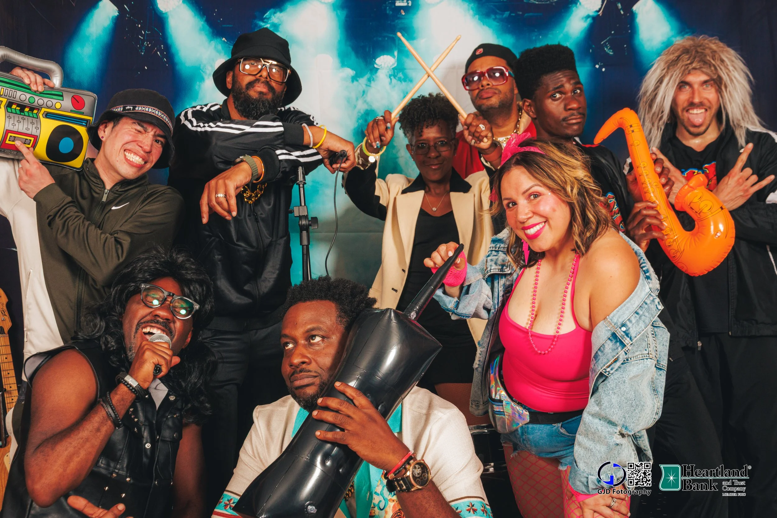 Group of people posing with humorous and retro costumes, including a woman holding a toy microphone, a man with a boombox, a man with drumsticks, and others with sunglasses, inflatable guitar, and eclectic outfits, in front of a colorful background.