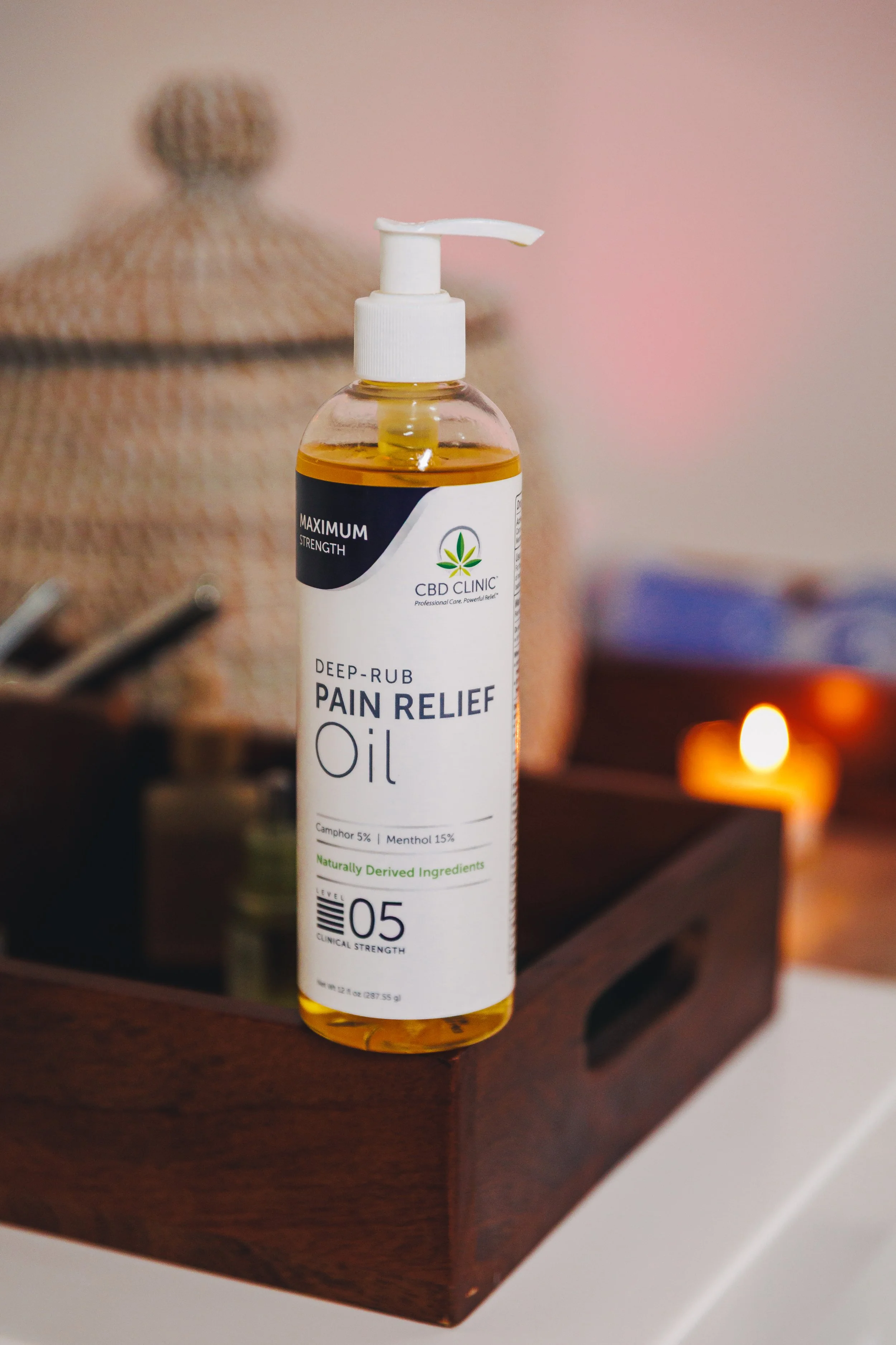 A bottle of CBD pain relief oil with a pump dispenser on a wooden tray next to a lit candle, and a woven basket in the background.