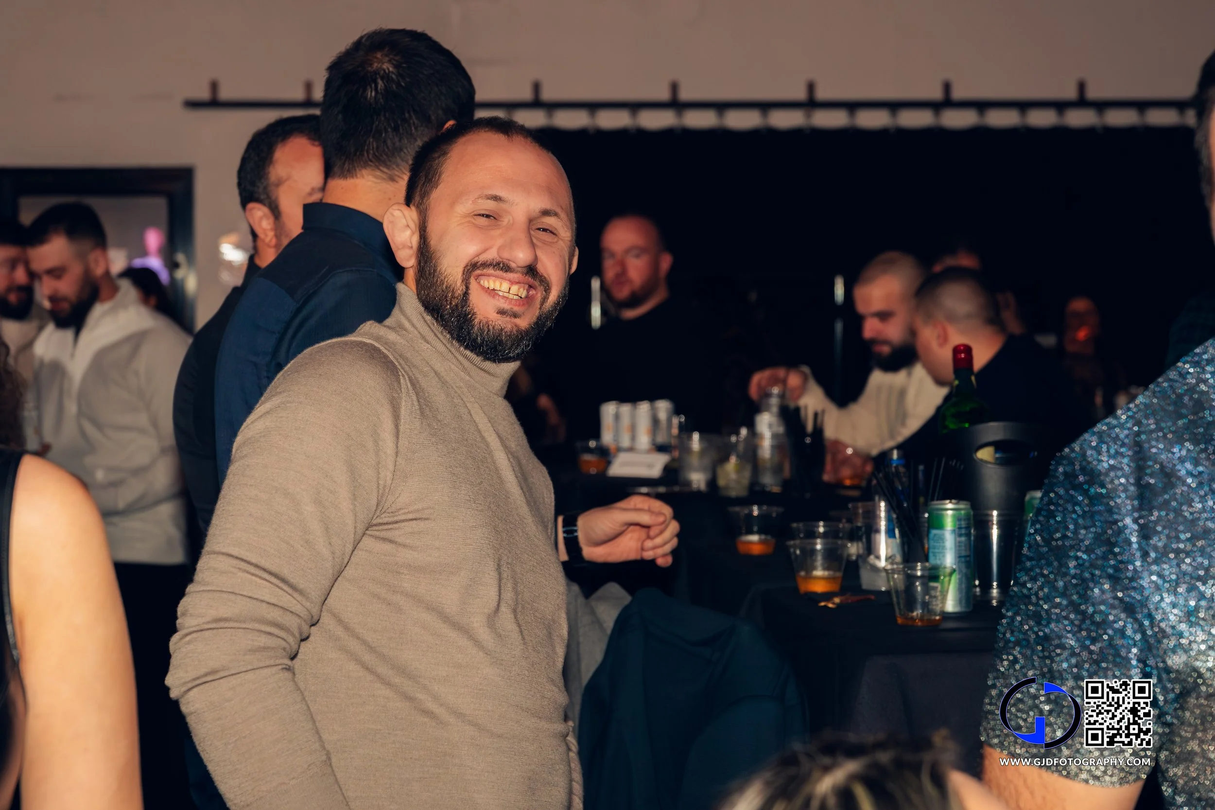 A smiling man with a beard in a beige turtleneck at a social gathering or party, with other people in the background near a bar.