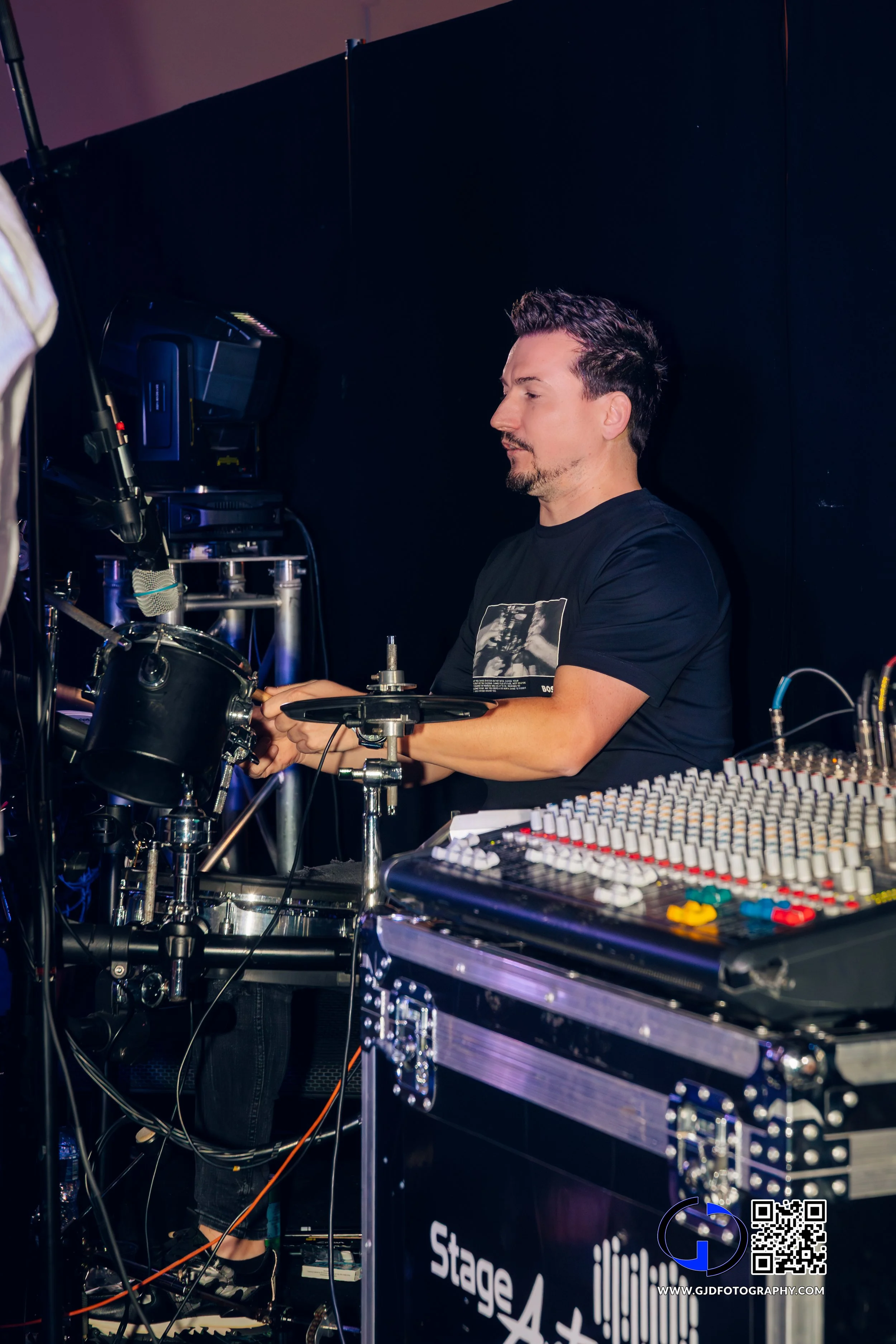 A male sound engineer adjusting sound equipment at a professional event or concert, with audio mixing console and DJ equipment visible.