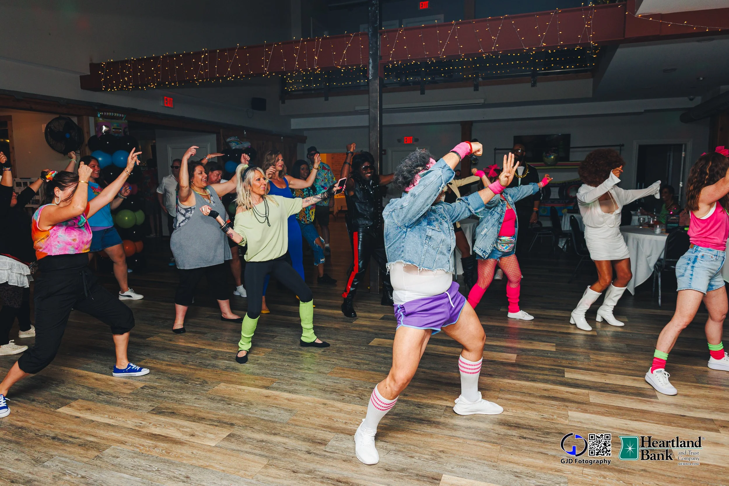 People dancing at a party with colorful clothing, neon accessories, and retro attire in a decorated venue with balloons and string lights.