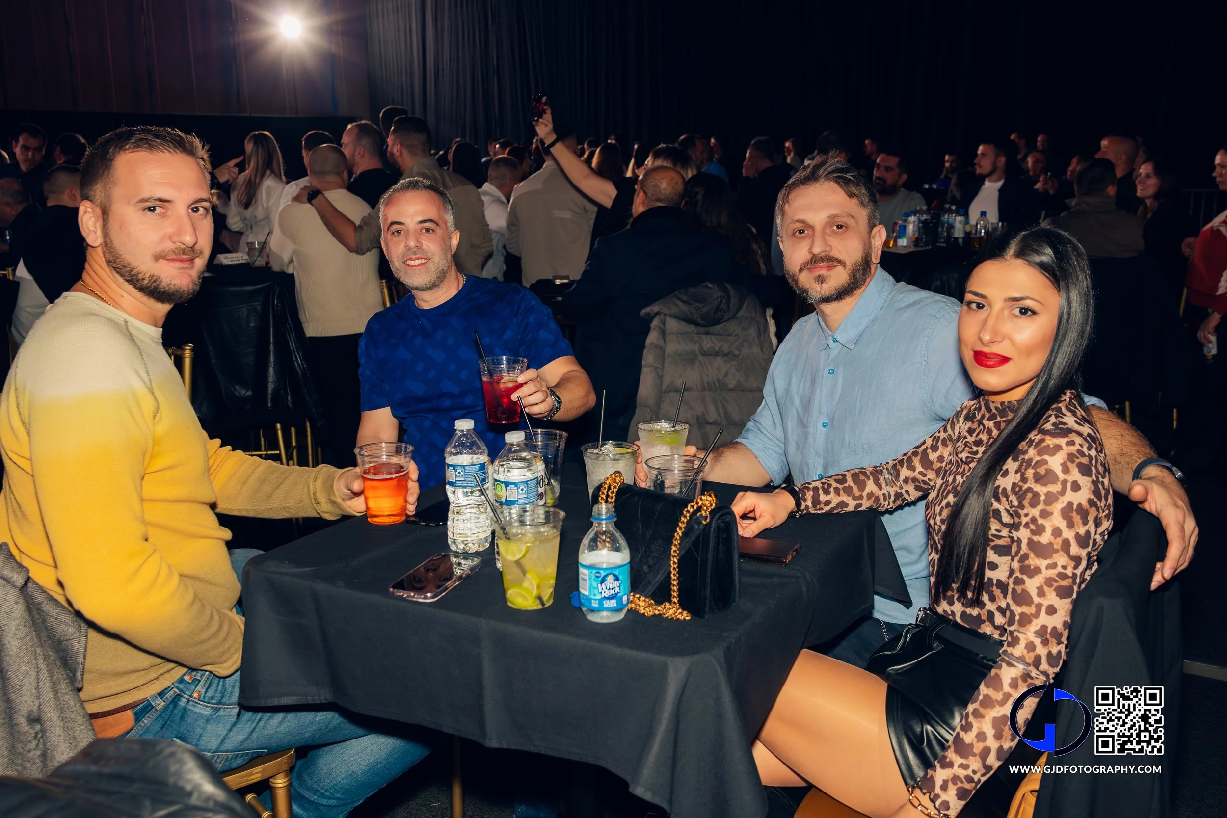 A group of five friends sitting at a table dressed casually, enjoying drinks at a crowded indoor event or party with a dark backdrop and a stage or screen in the background.