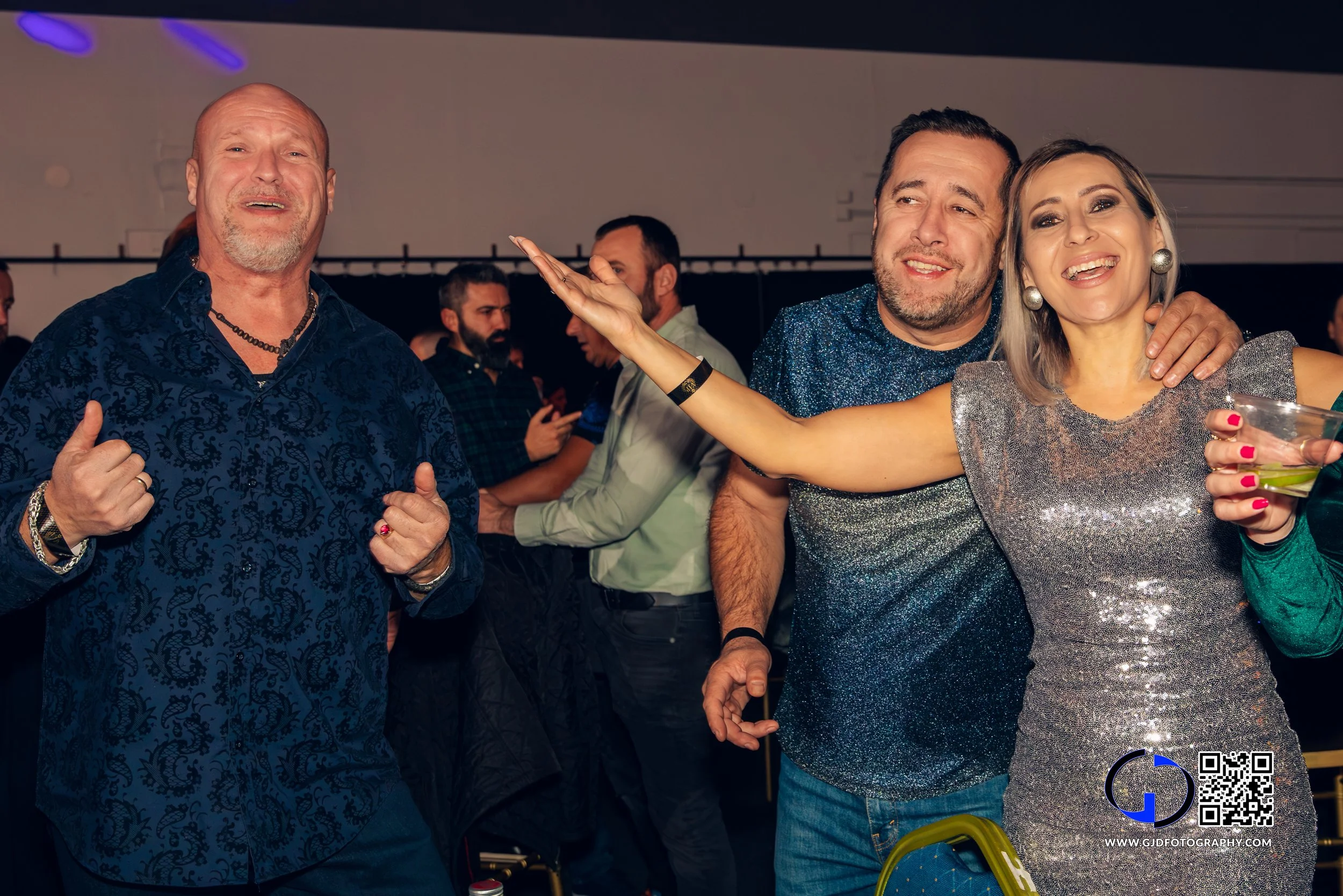 People dancing and socializing at a party, smiling and enjoying themselves, with a man in a blue patterned shirt, a woman in a sparkly dress, and another man in a blue shirt.