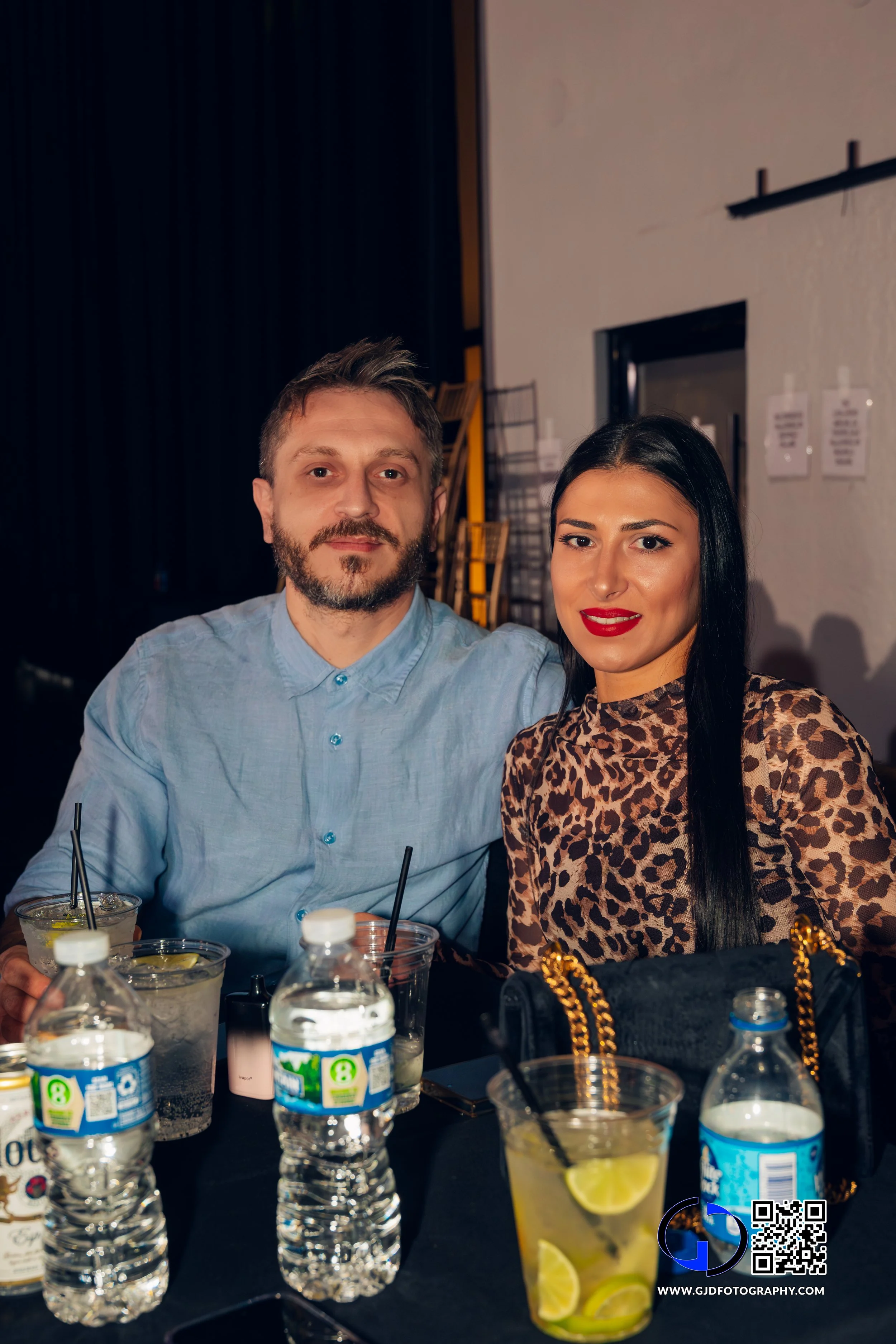 A man with short brown hair and beard, wearing a light blue button-up shirt, and a woman with long black hair, wearing a leopard print top, sitting together at a table with drinks at a social gathering or restaurant.