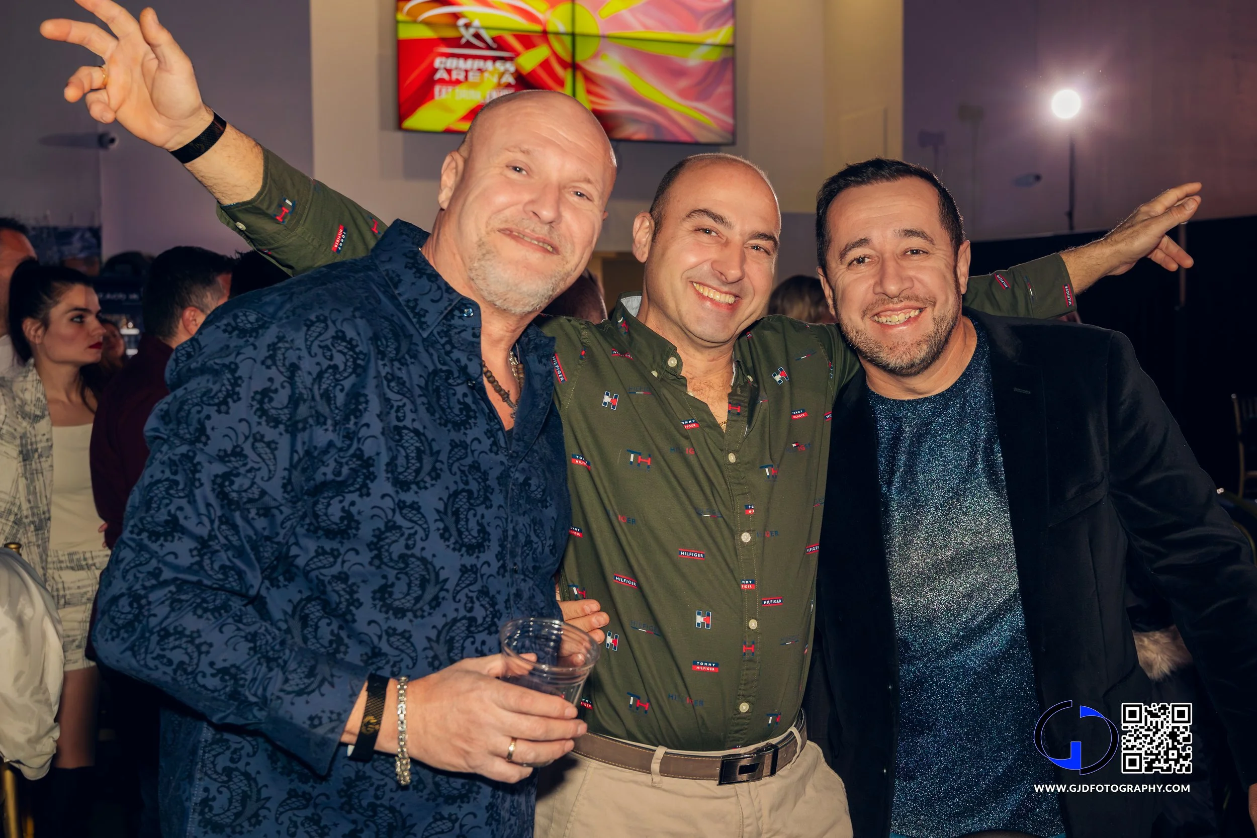 Three men smiling and posing together at a party or event. The man on the left is holding a drink, the man in the middle has arms around the others, and the man on the right is wearing a black jacket.