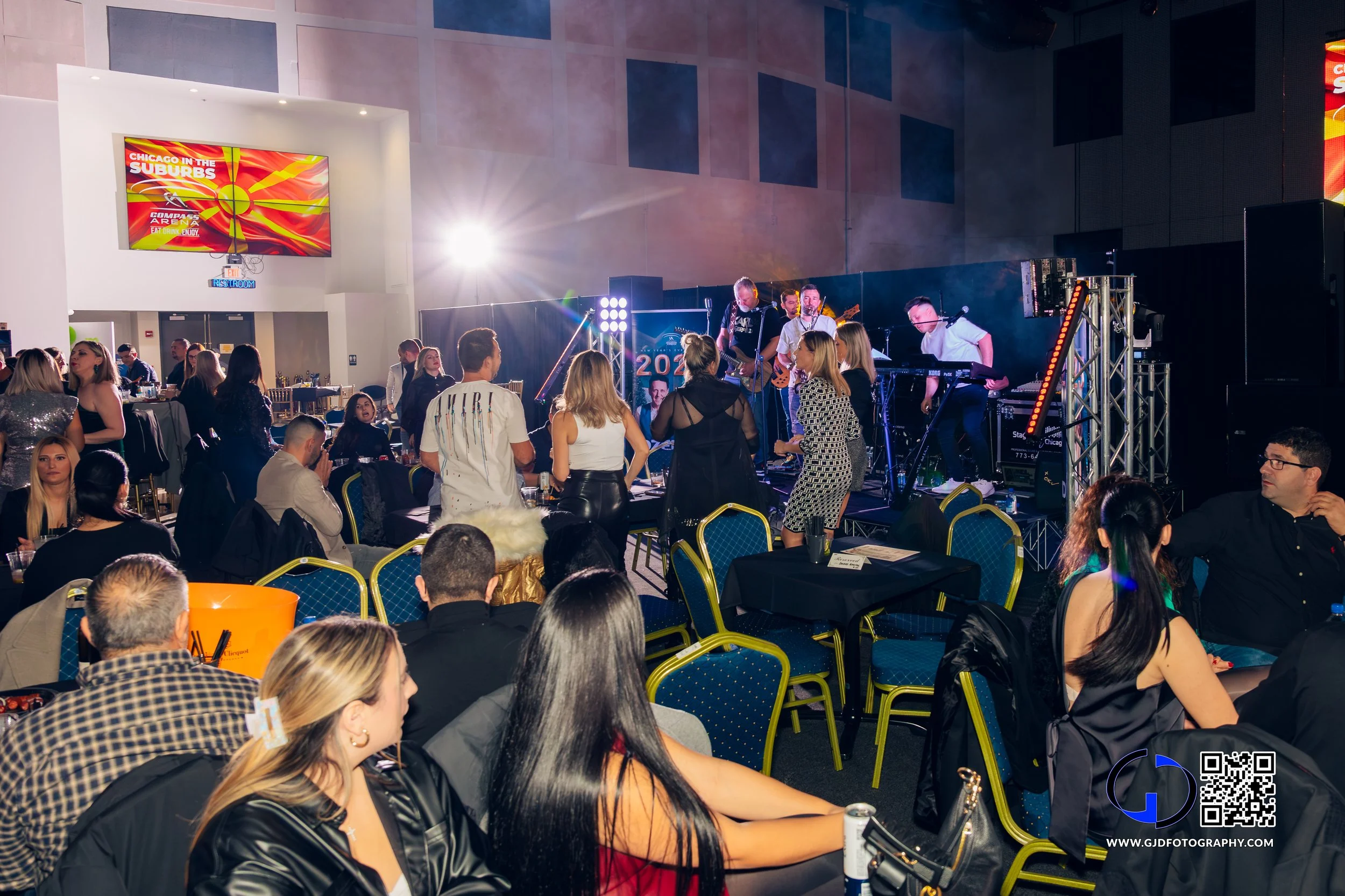 Indoor concert event with people watching a live band on stage, with some standing and dancing, guests seated at tables, and a large digital screen displaying event info in the background.