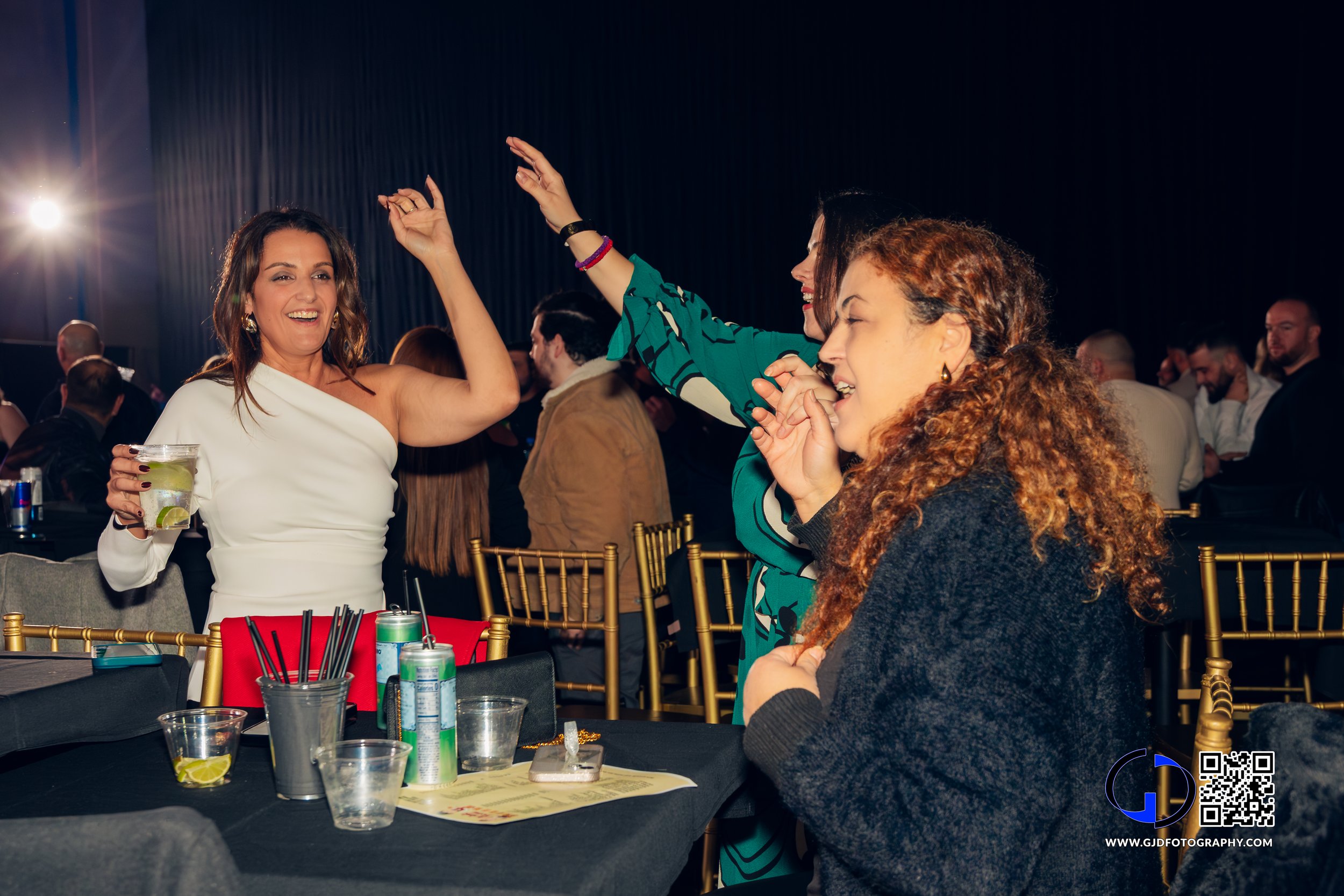 Three women enjoying a night out at a party, dancing and smiling, with drinks on the table and a crowd of people in the background.