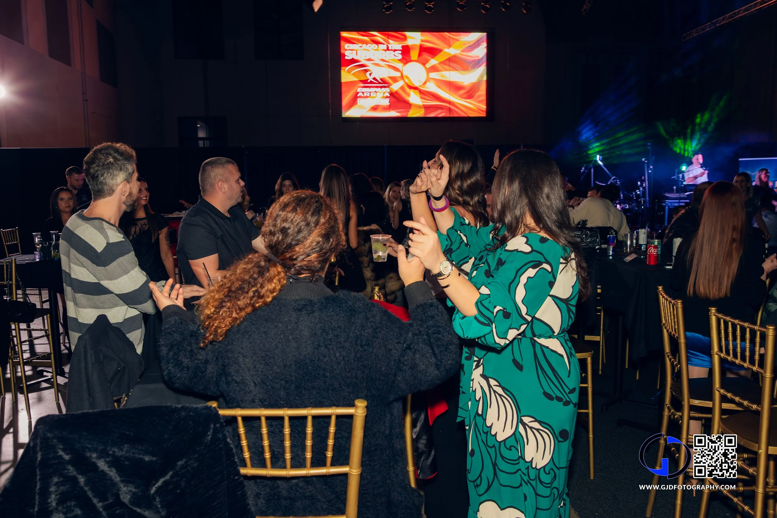People dancing and socializing at a concert or event with a large LED screen and stage with musicians in the background.