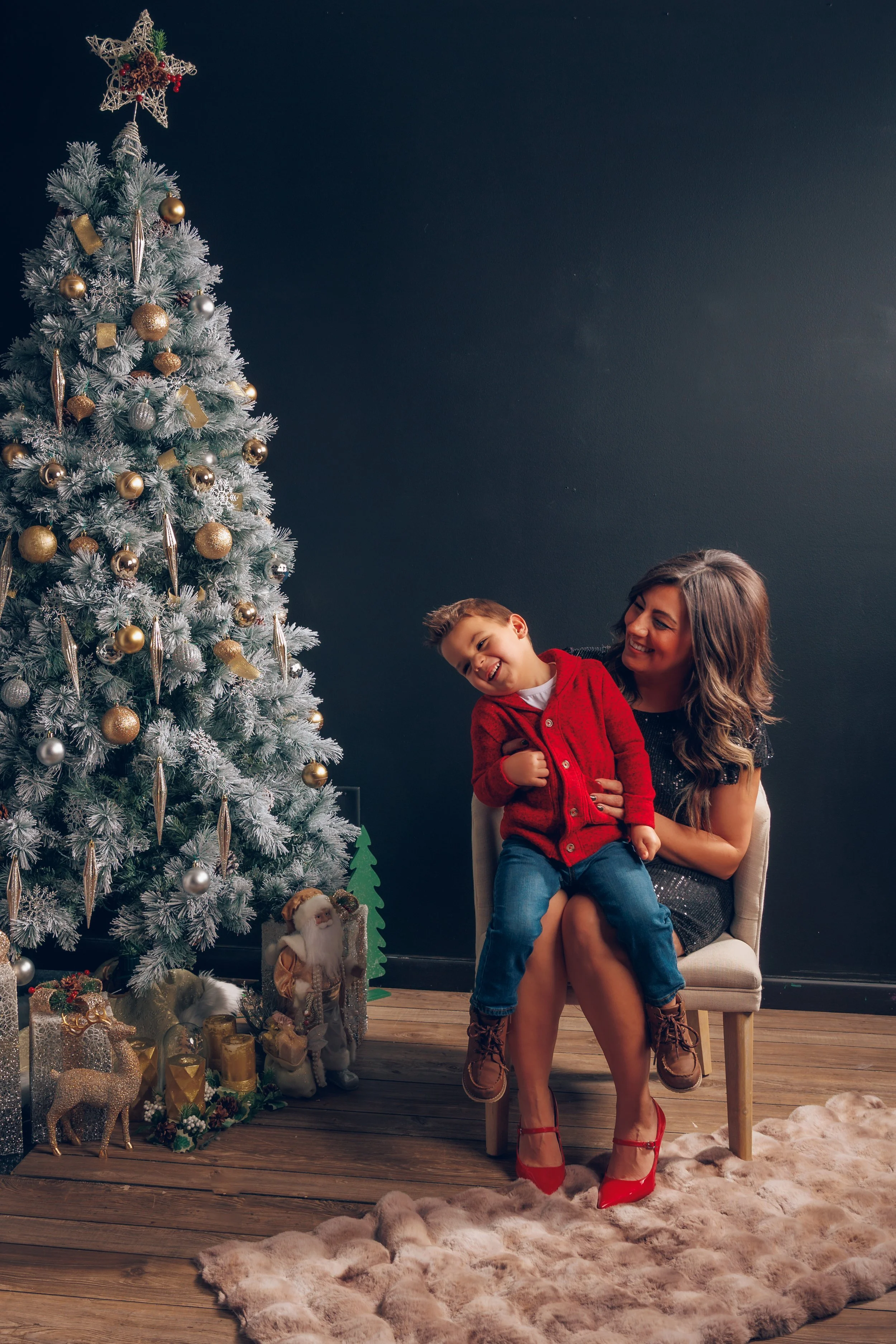 A woman and a boy sitting on a chair next to a decorated Christmas tree with ornaments, gifts, and holiday decorations.