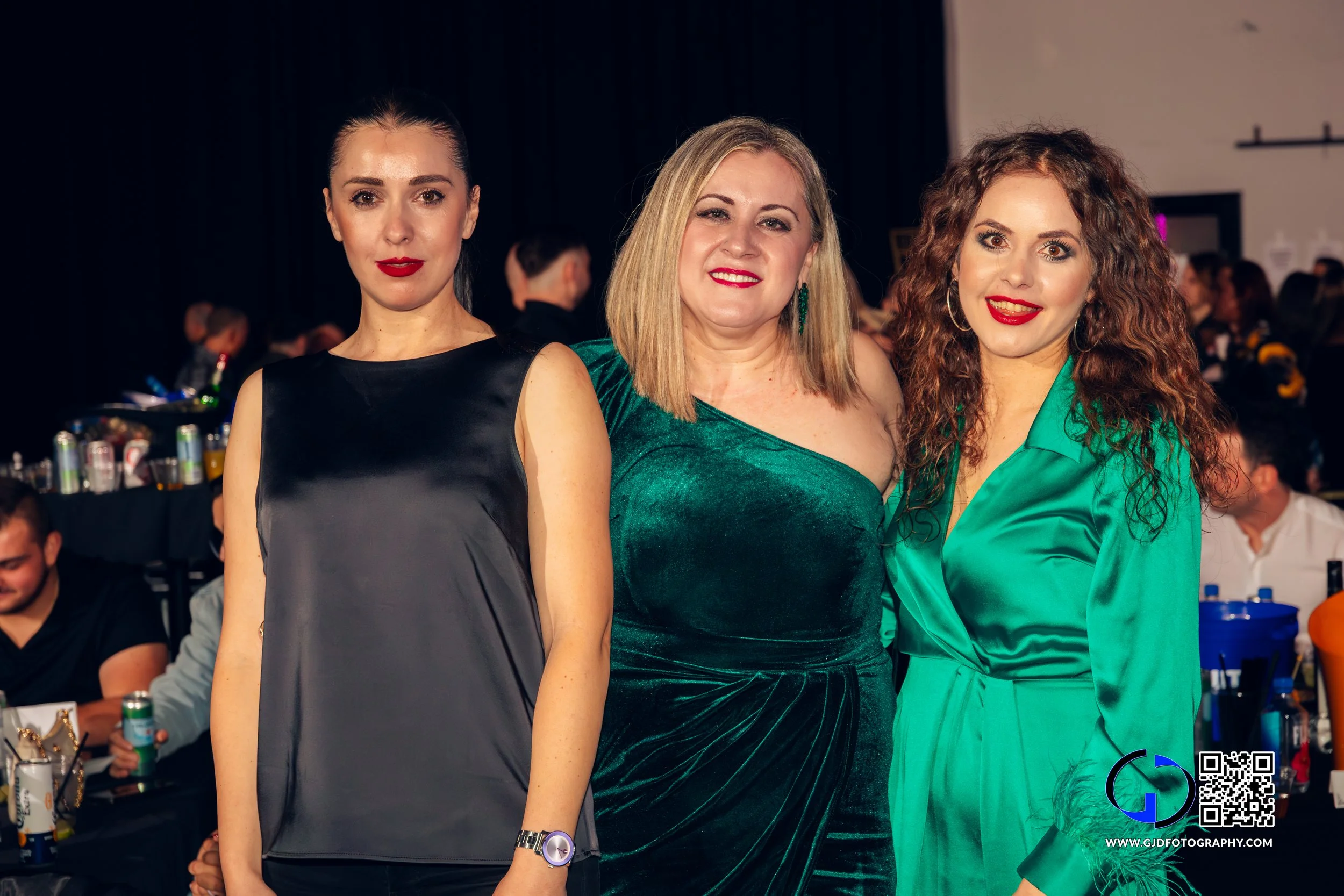 Three women standing together at an indoor event, smiling at the camera. The woman on the left has dark hair styled in a high ponytail, wearing a sleeveless black and gray top, and a watch. The woman in the middle has blonde hair, wearing a green off