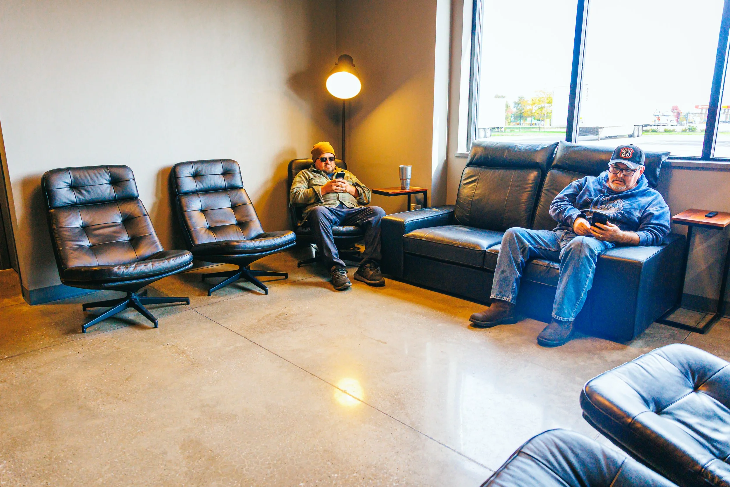 Two men sitting on black leather sofas and chairs in a waiting room, using their smartphones. The room has a large window, a standing lamp, and a side table.