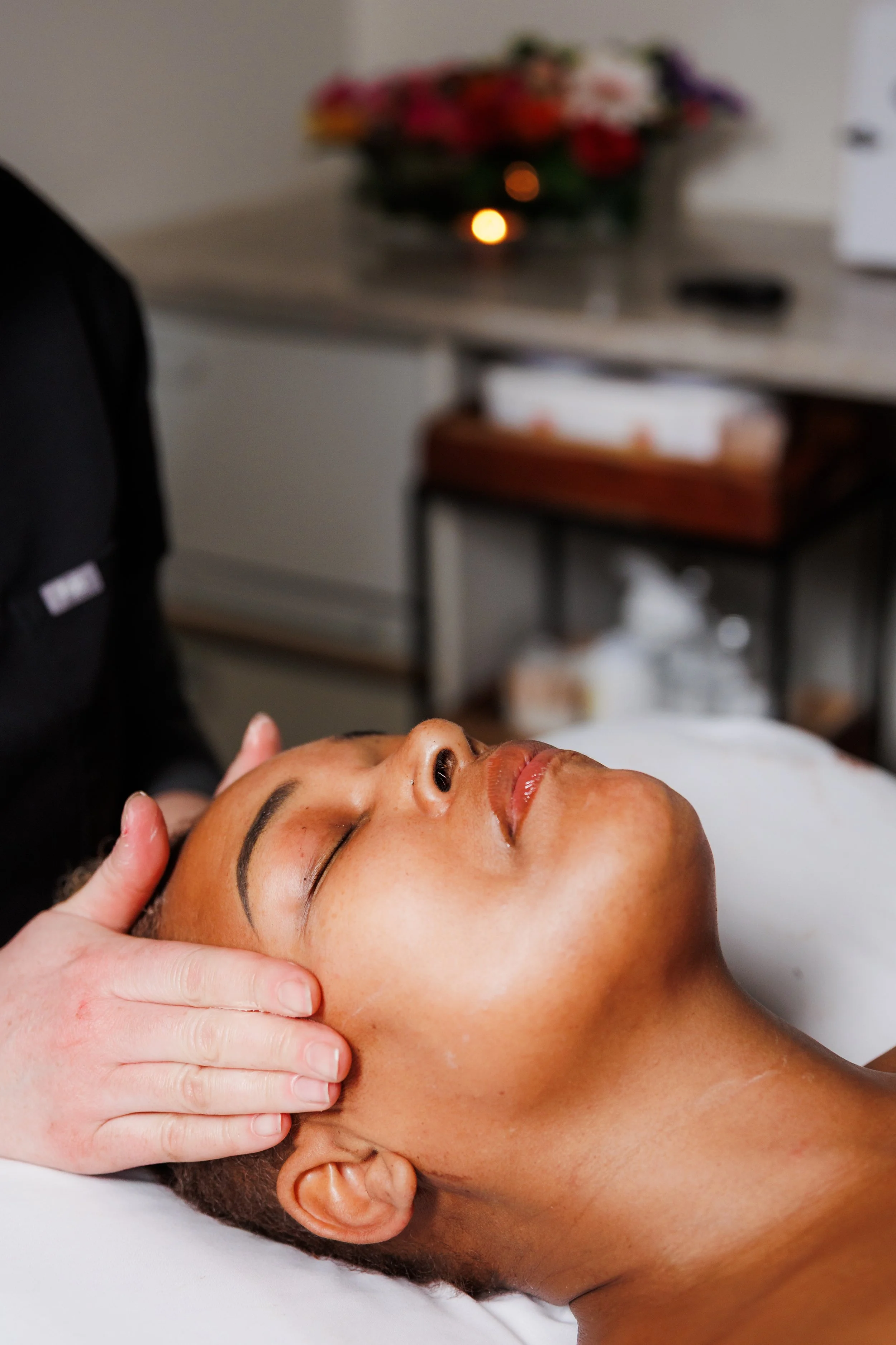 A woman receiving a massage or facial treatment in a spa or clinic, with a bouquet of flowers and a candle in the background.