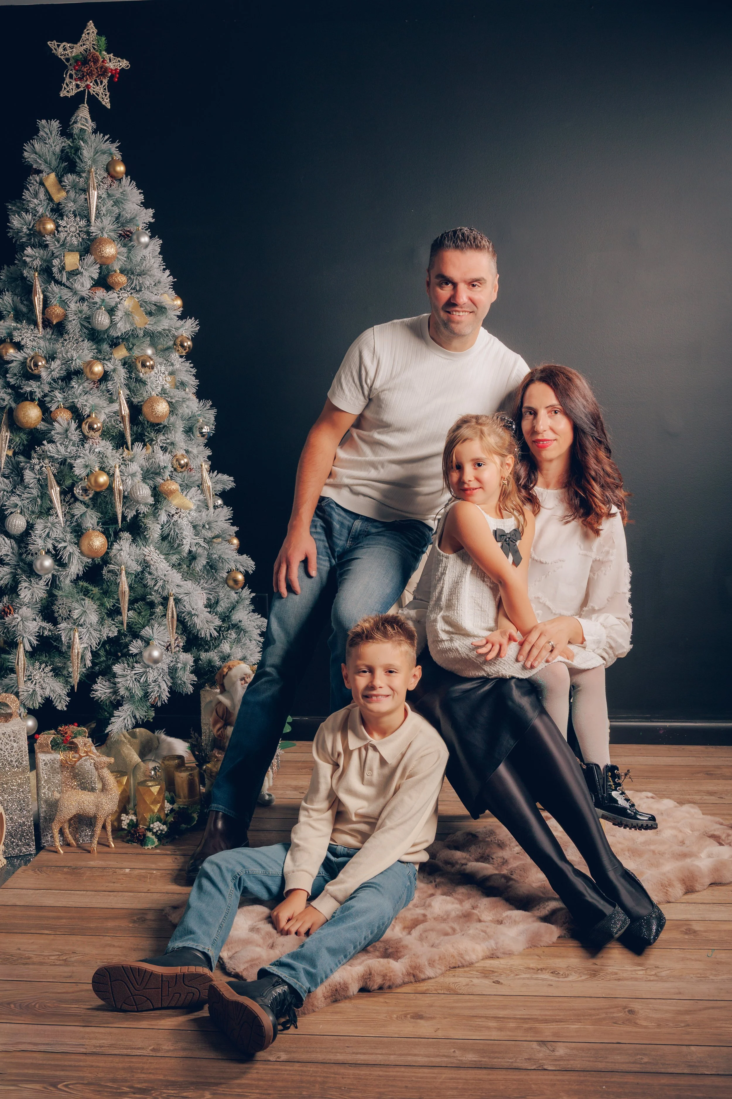 A family of four sitting and standing around a Christmas tree decorated with gold and silver ornaments, with wrapped gifts underneath, in a cozy indoor setting.