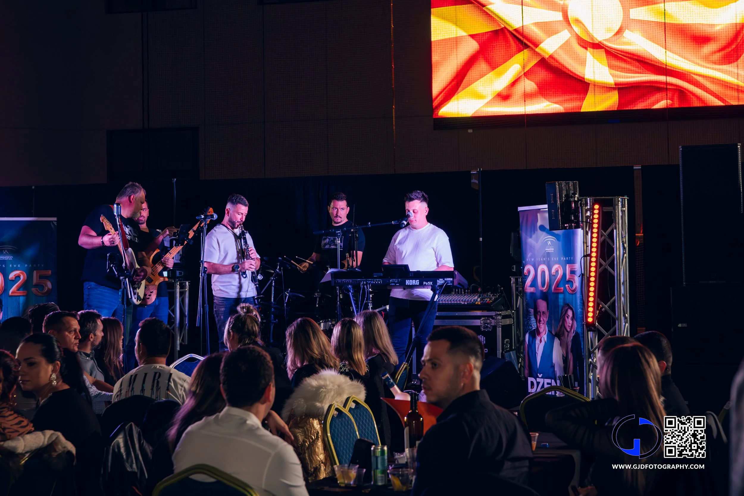 Musicians performing on stage at an event with an audience seated at round tables, a large digital screen displaying a bright abstract design, and promotional banners with the year 2025.