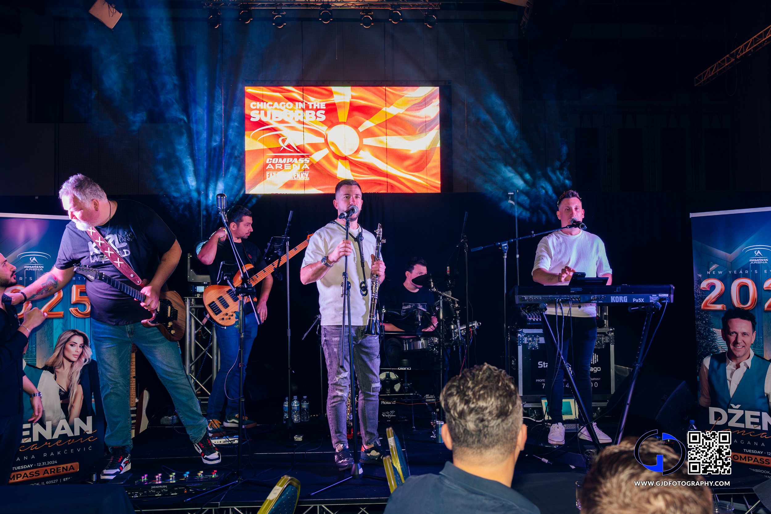 A band performs on stage with keyboards, guitars, and drums at a concert in an arena, with a large screen in the background displaying event information.